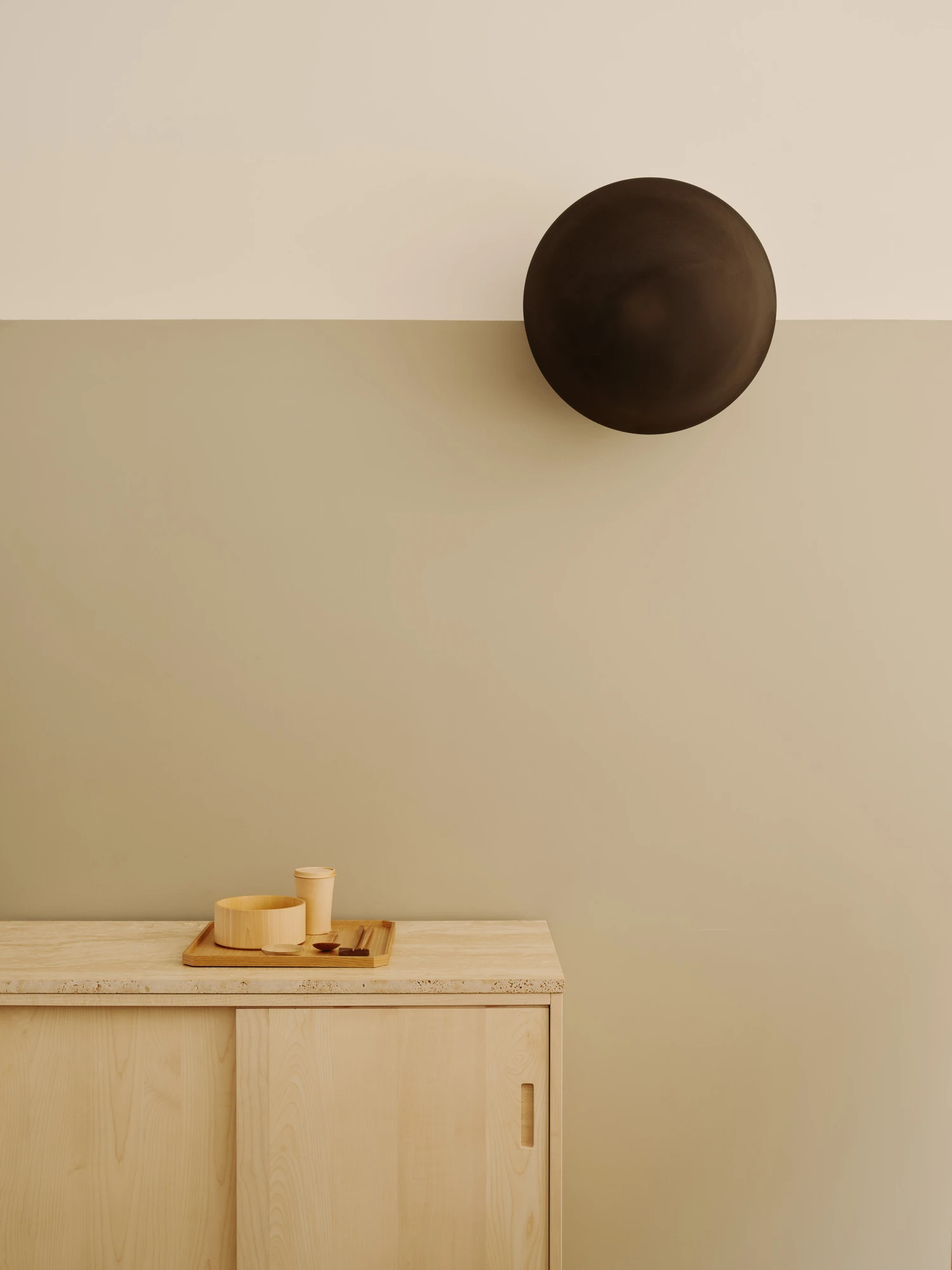 Nichigetsu by Studio Inhalt_, photographed by Robert Rieger — A dark bronze disc light sits on the seam of a cream-and-sage wall above a maple cabinet topped in travertine, a tray of pale vessels below.