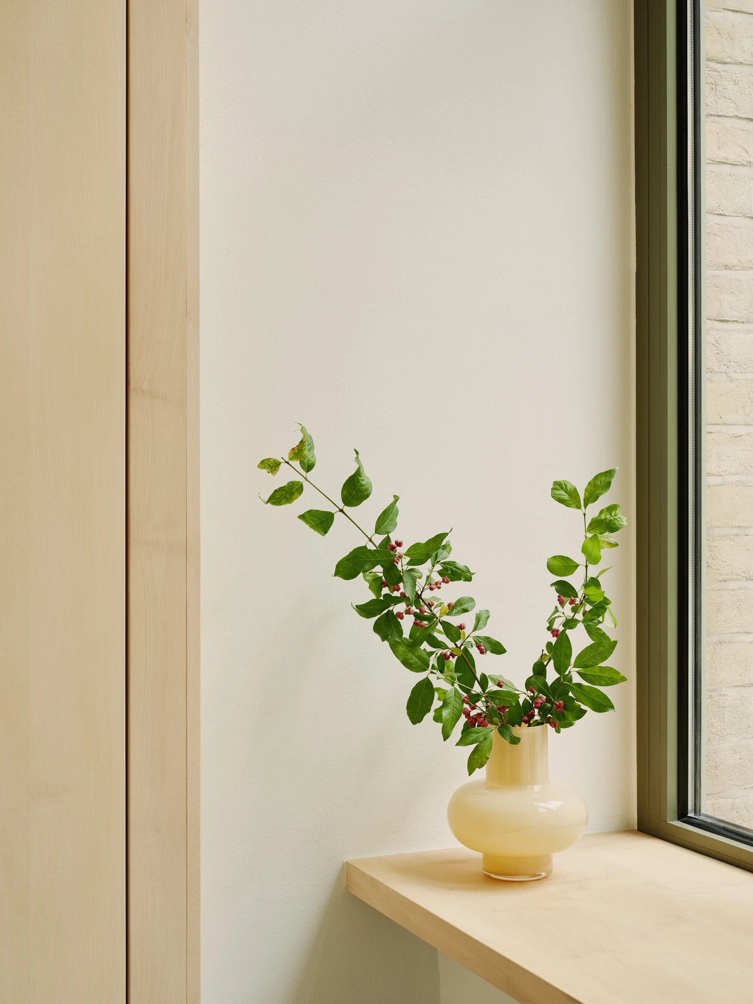Nichigetsu by Studio Inhalt_, photographed by Robert Rieger — A pale maple sill holds a cream glass vase of leafed branches and red berries against an olive-framed window and brick beyond.