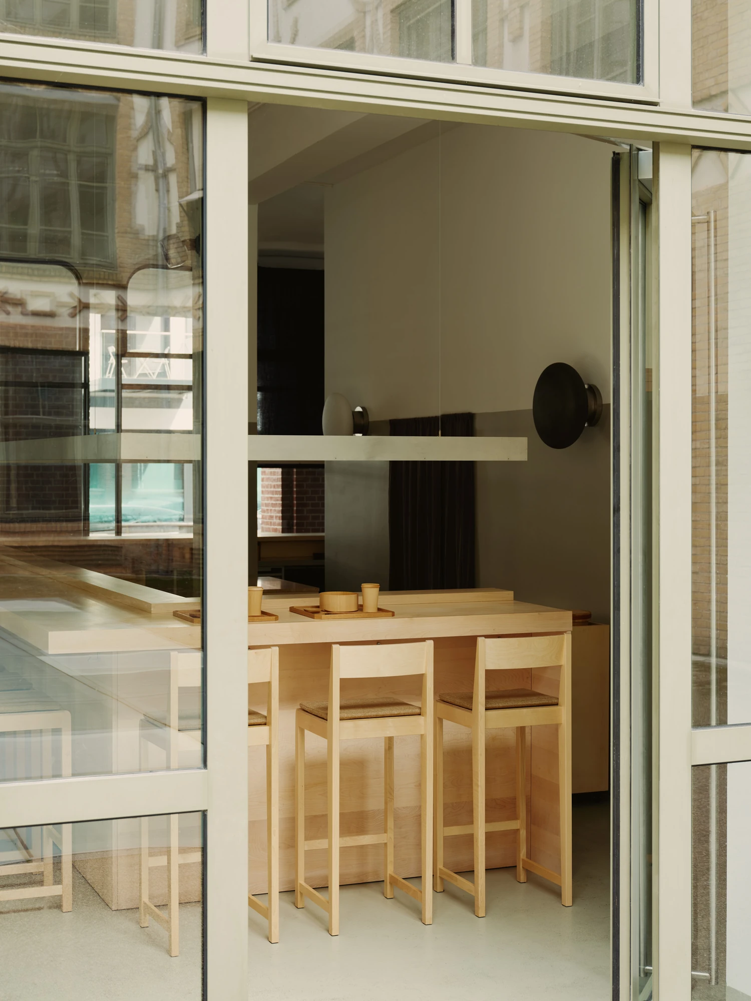 Nichigetsu by Studio Inhalt_, photographed by Robert Rieger — Seen through the glazed facade, the maple counter and its tall stools sit under a bronze disc light, street reflections crossing the glass.