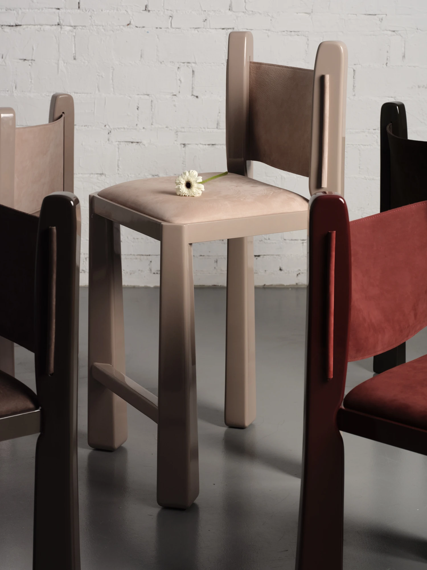 Ruby by NOVSEP, photographed by NOVSEP — A cluster of Ruby counter stools in oxblood, greige and espresso high-gloss lacquer gathers against a whitewashed brick wall, a single white gerbera resting on one suede seat.