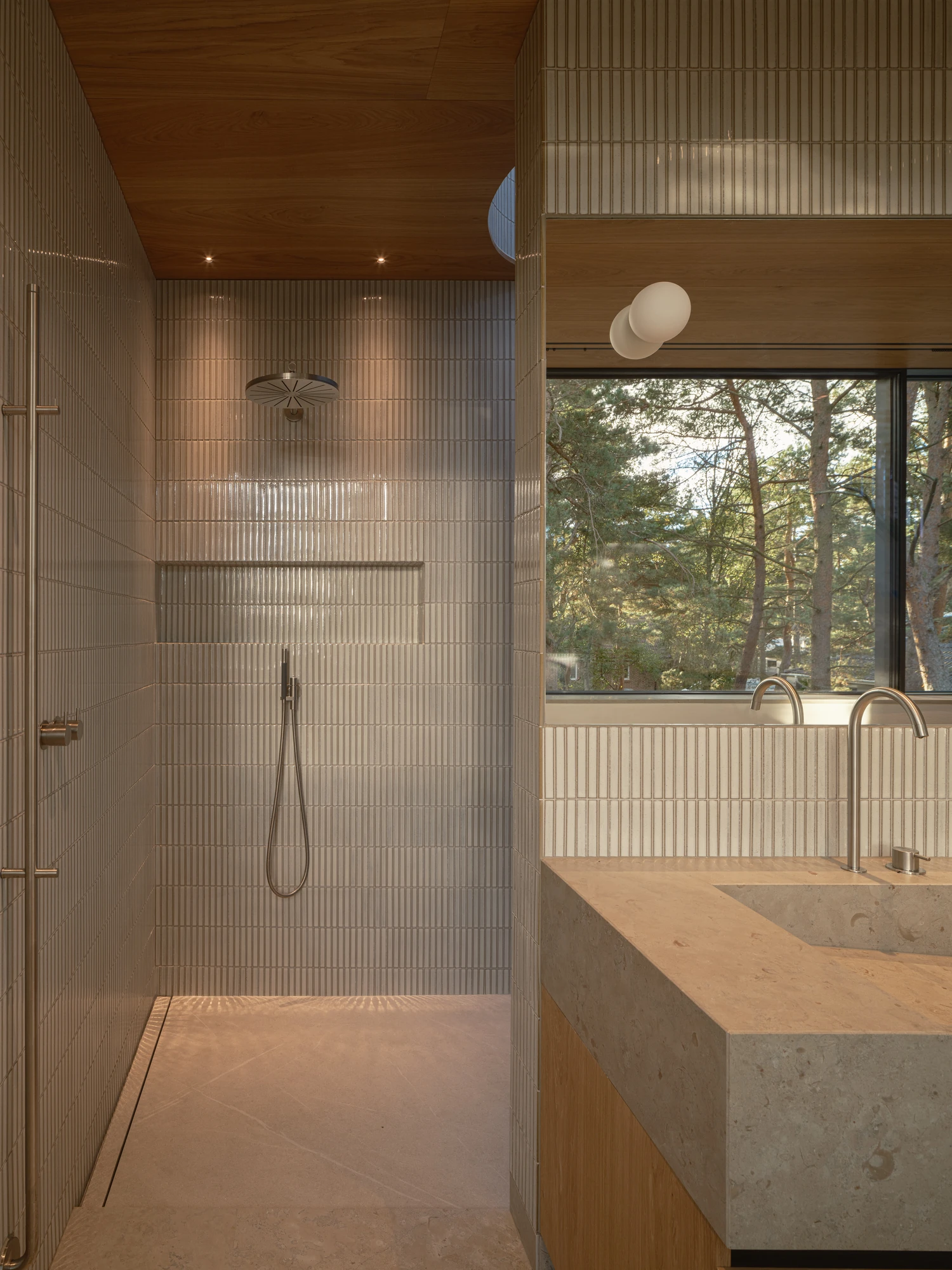 Villa P by Daniel Berg Arkitekter, photographed by Markus Linderoth — Off-white finger tiles wrap a walk-in shower next to a limestone basin set below a window framing the surrounding trees.