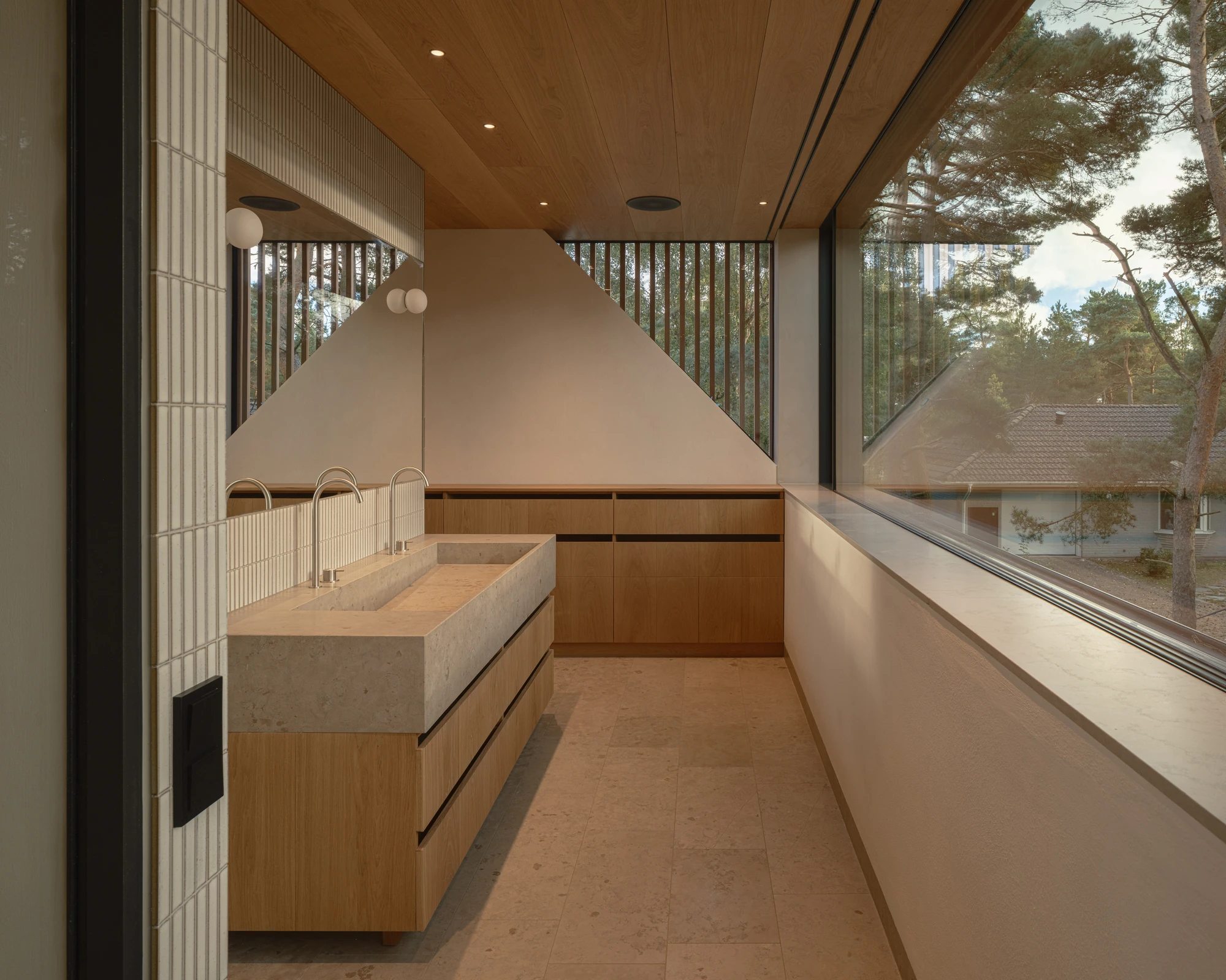 Villa P by Daniel Berg Arkitekter, photographed by Markus Linderoth — A stone trough sink runs beneath a tall mirror in an oak-lined bathroom, floor-to-ceiling glazing opening the room to the pines.