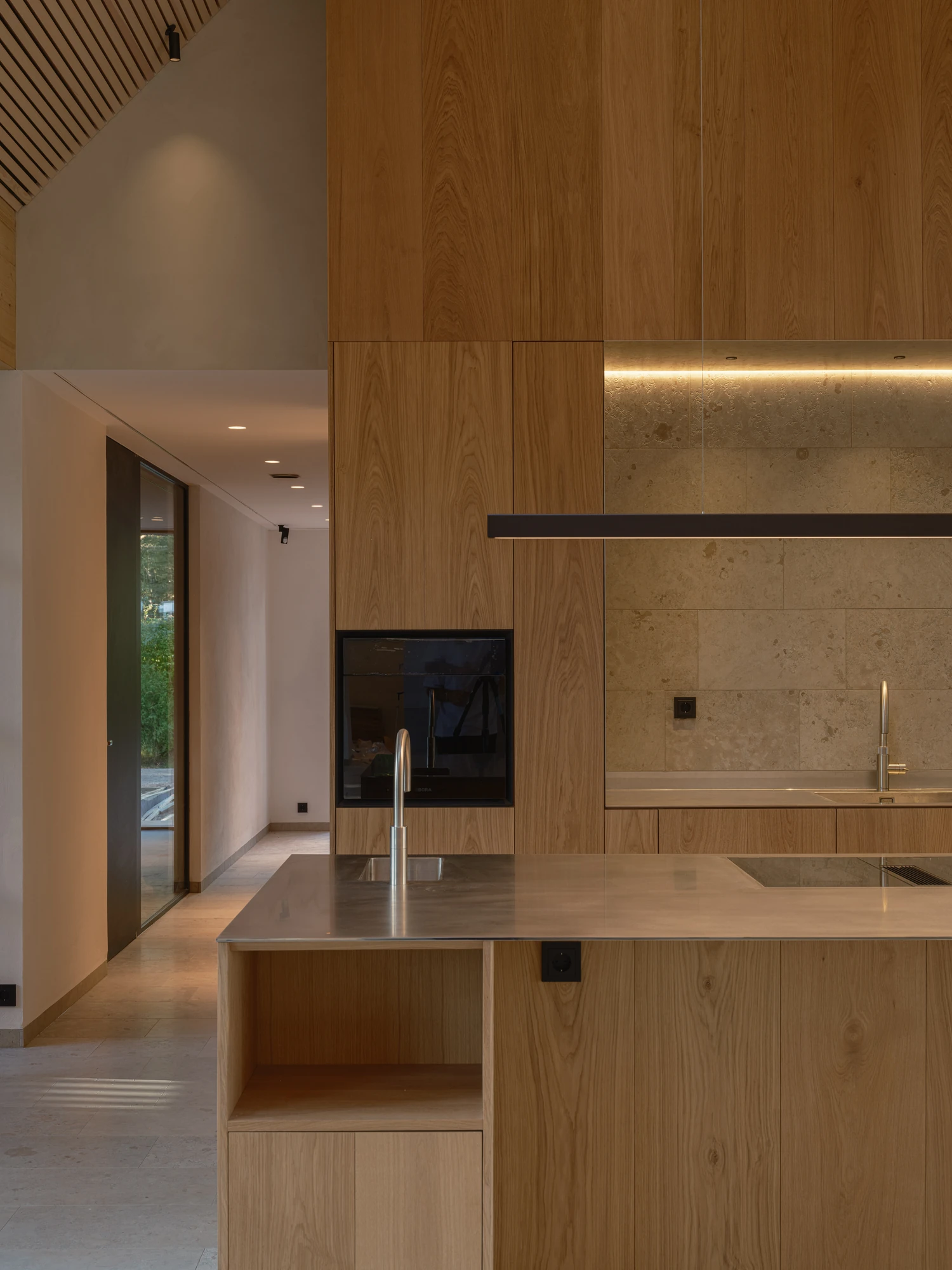 Villa P by Daniel Berg Arkitekter, photographed by Markus Linderoth — A stainless steel island stands against oak cabinetry and a limestone backsplash, a glazed corridor drawing daylight from the far end.