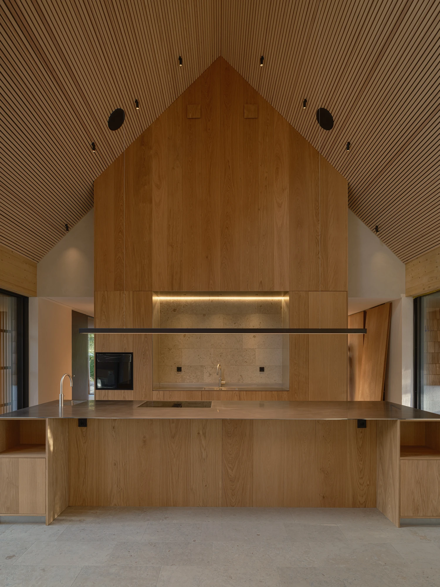 Villa P by Daniel Berg Arkitekter, photographed by Markus Linderoth — Under a gable lined with oak slats, an oak-clad volume rises above a stainless island and limestone splashback on a limestone floor.
