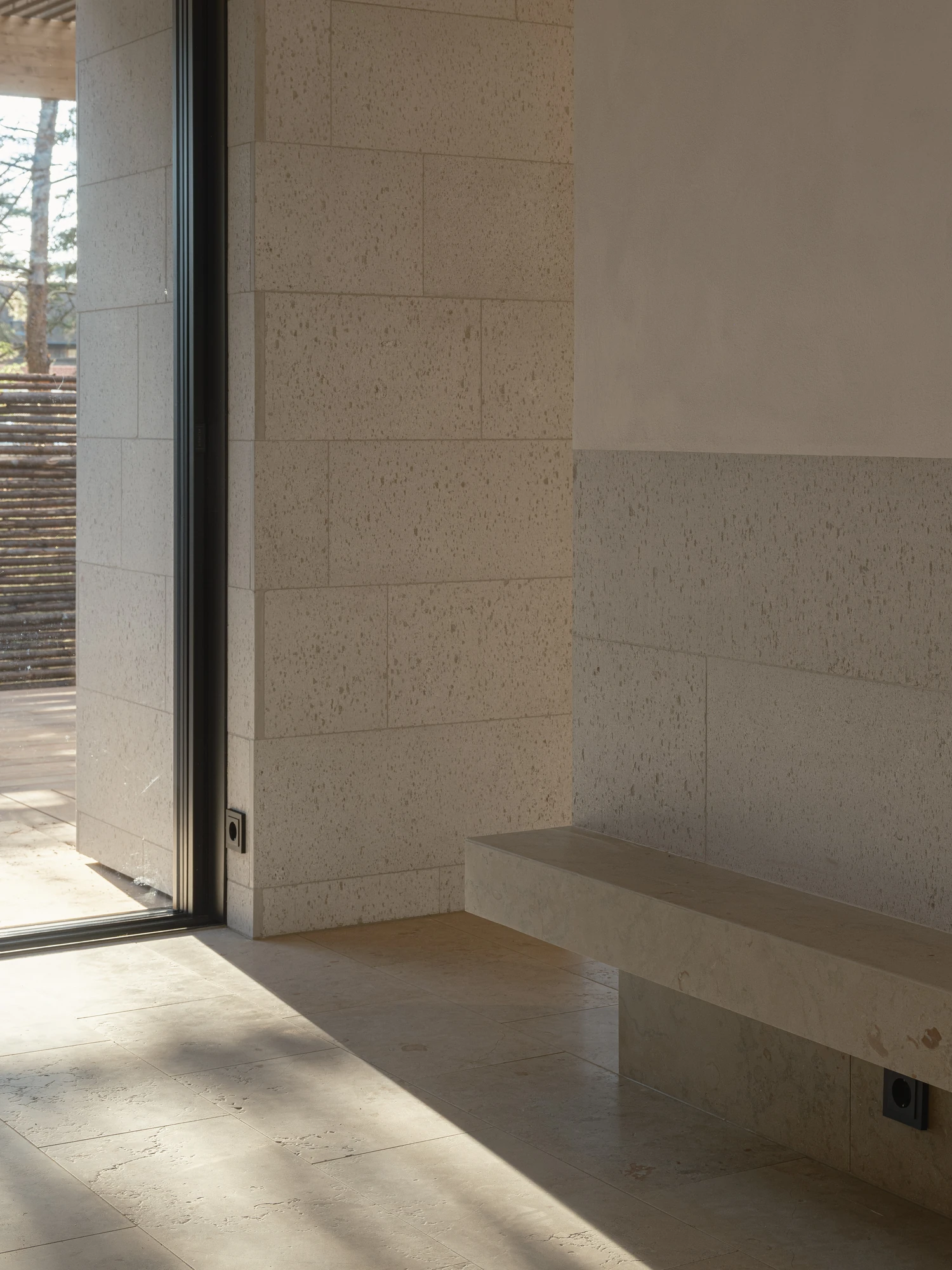 Villa P by Daniel Berg Arkitekter, photographed by Markus Linderoth — Sunlight falls across a limestone floor and a cantilevered stone bench beside floor-to-ceiling glazing set into a pale limestone wall.