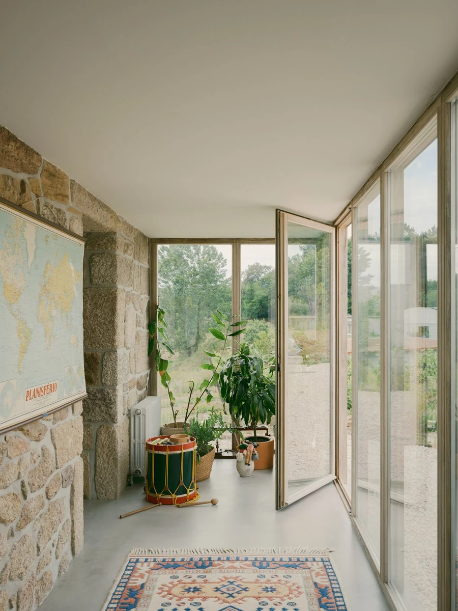 Casa de Souvinha by Parto Atelier, photographed by Francisco Ascensão, Parto Atelier — A glazed veranda floored in pale microcement runs along an exposed granite wall hung with a world map, a kilim rug and painted drum grounding the corner of light.