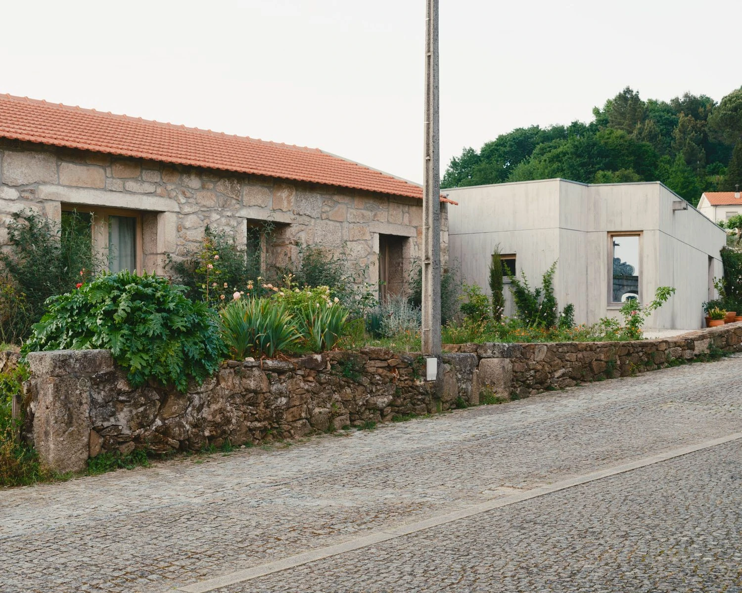 Casa de Souvinha by Parto Atelier, photographed by Francisco Ascensão, Parto Atelier — A red-tiled granite cottage sits along the cobbled village road, the new concrete volume reading as its quiet neighbour beyond a flower border.