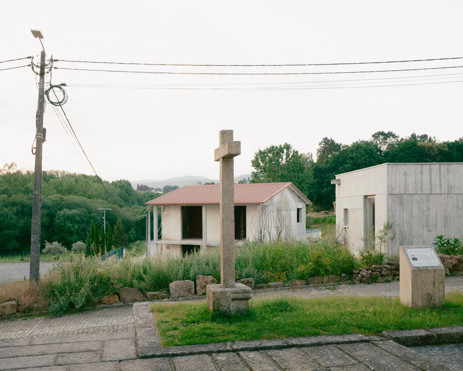 Casa de Souvinha by Parto Atelier, photographed by Francisco Ascensão, Parto Atelier — A weathered granite cross stands in the foreground as the concrete home and an older tiled house share the sloping village ground under an overcast sky.