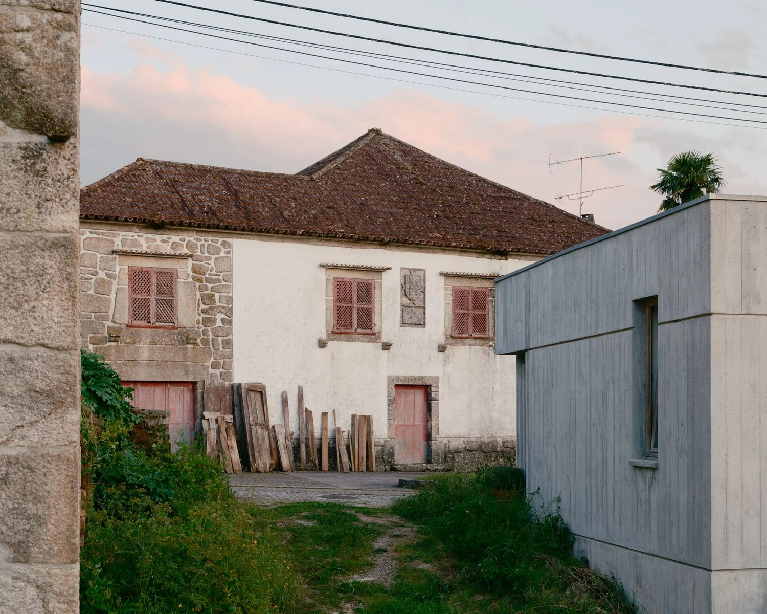 Casa de Souvinha by Parto Atelier, photographed by Francisco Ascensão, Parto Atelier — An old rendered manor with a moss-darkened tile roof and rust-red shutters faces the smooth grey flank of the concrete volume across a grassy yard at dusk.