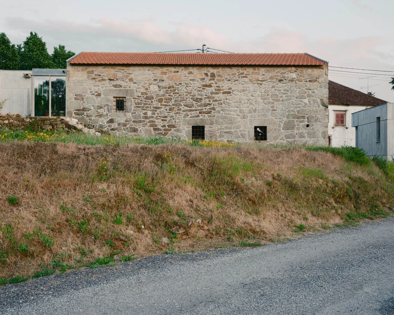 Casa de Souvinha by Parto Atelier, photographed by Francisco Ascensão, Parto Atelier — The long granite wine cellar carries a red-tiled roof above a grassy embankment, the glazed link and concrete home visible at its far end along the gravel road.