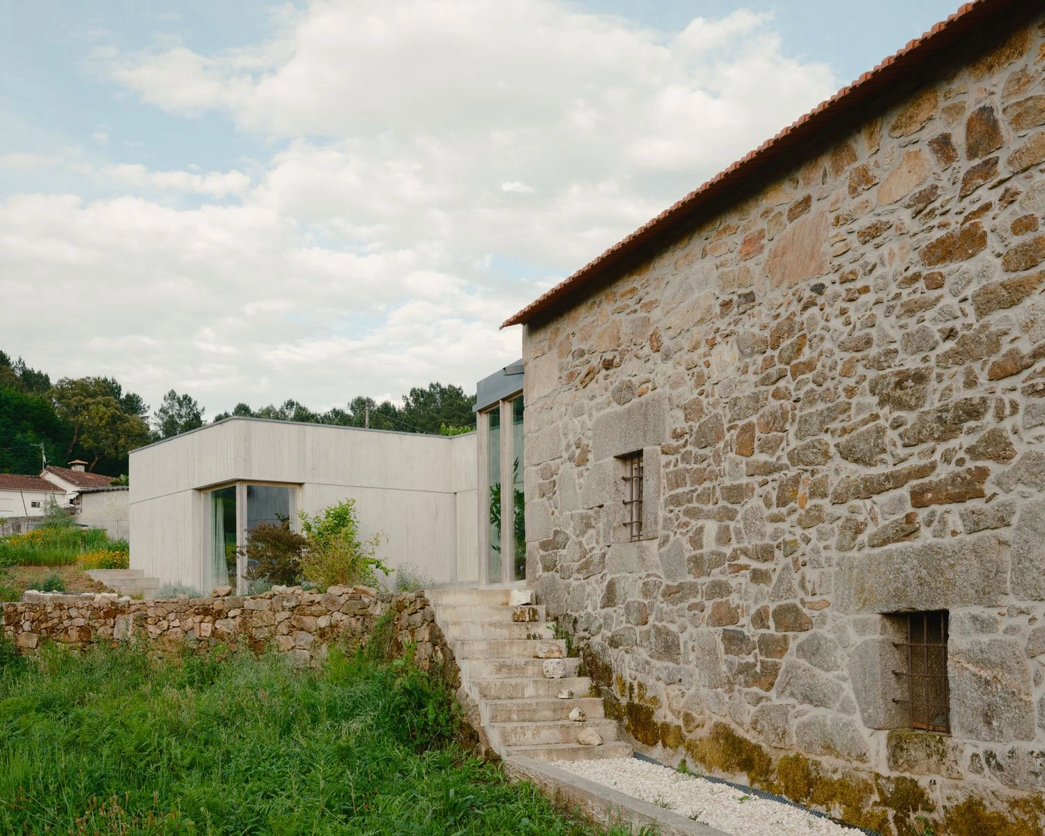 Casa de Souvinha by Parto Atelier, photographed by Francisco Ascensão, Parto Atelier — Cast concrete steps climb between the granite masonry gable and the new volume, the glazed link opening the garden behind a tangle of grass.