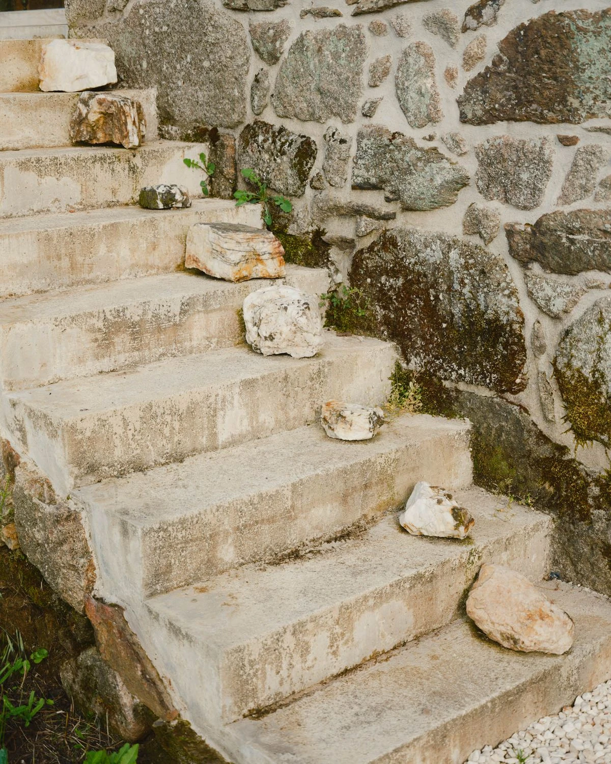 Casa de Souvinha by Parto Atelier, photographed by Francisco Ascensão, Parto Atelier — White quartz stones rest on rough cast concrete steps beside a moss-flecked granite wall, a small deliberate gesture along the approach.