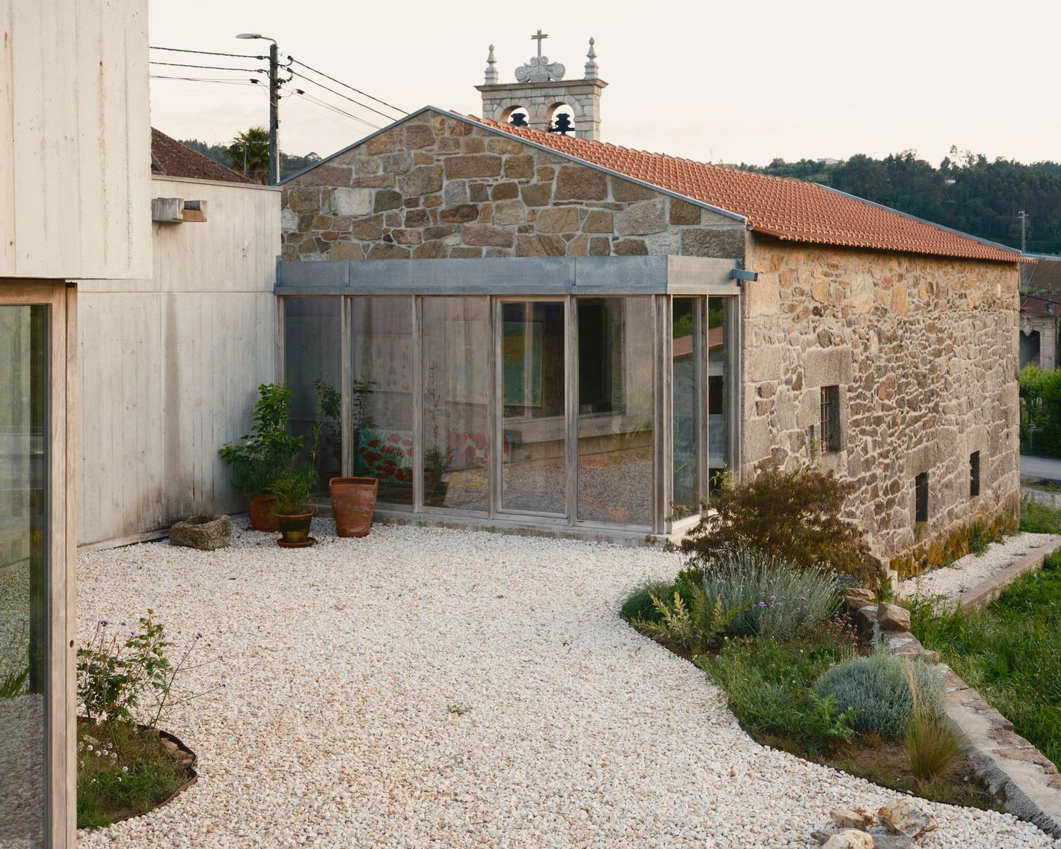 Casa de Souvinha by Parto Atelier, photographed by Francisco Ascensão, Parto Atelier — A pale gravel courtyard opens between the concrete home and the granite building, a zinc-capped glazed veranda between them and a church bell tower rising behind.