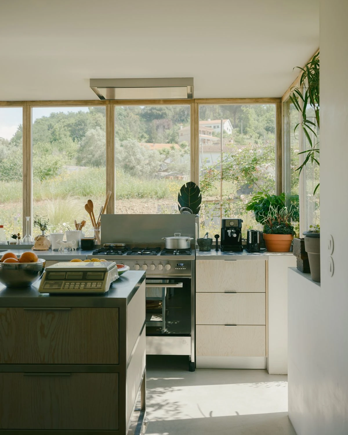 Casa de Souvinha by Parto Atelier, photographed by Francisco Ascensão, Parto Atelier — The kitchen sets a stainless steel counter and hood against blond pine cabinets, its corner of glazing framing the meadow and distant hillside houses.