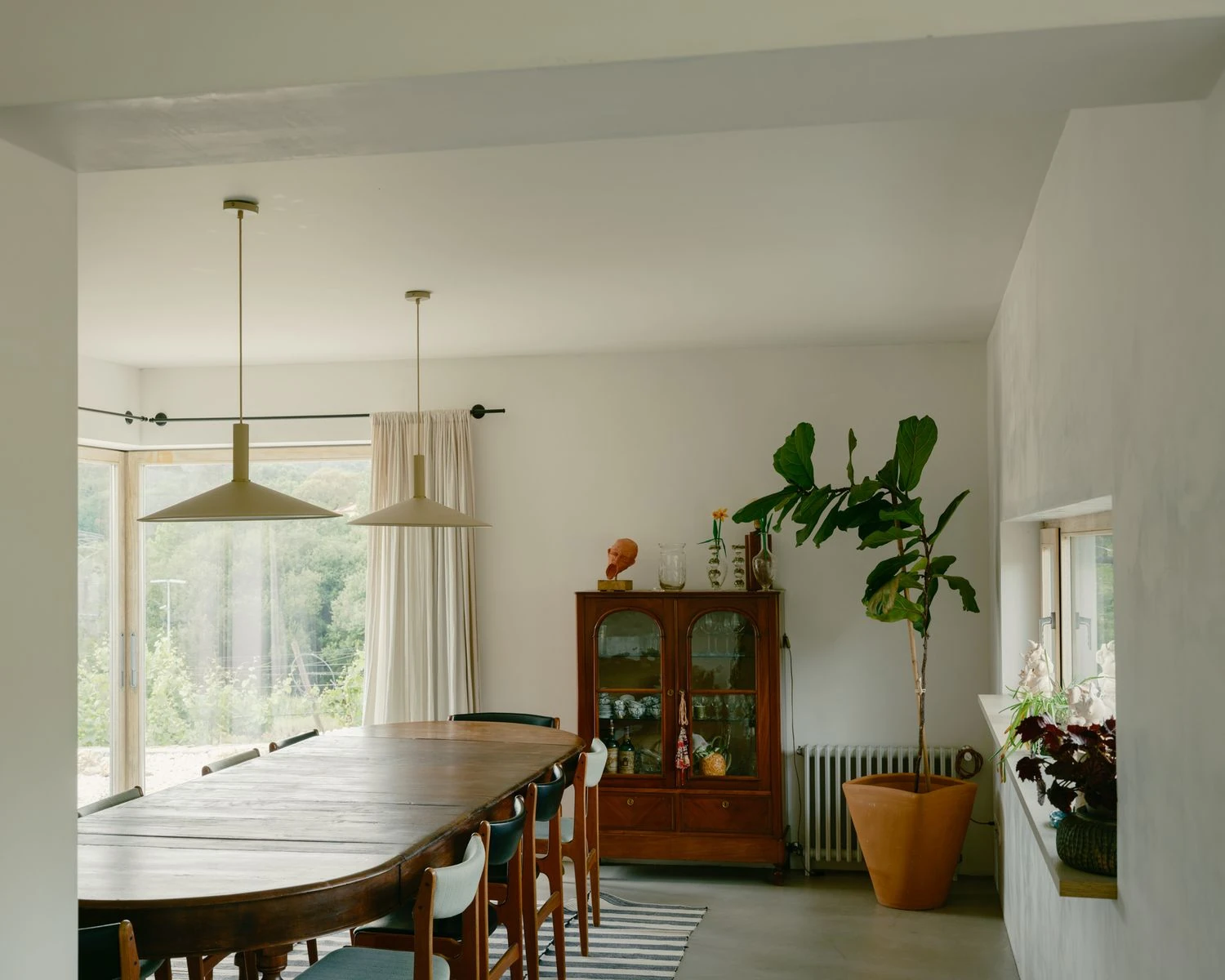 Casa de Souvinha by Parto Atelier, photographed by Francisco Ascensão, Parto Atelier — A long dark-wood table runs under two pendant lamps in the dining room, a glazed door opening to the garden beside an arched display cabinet and a fiddle-leaf fig.