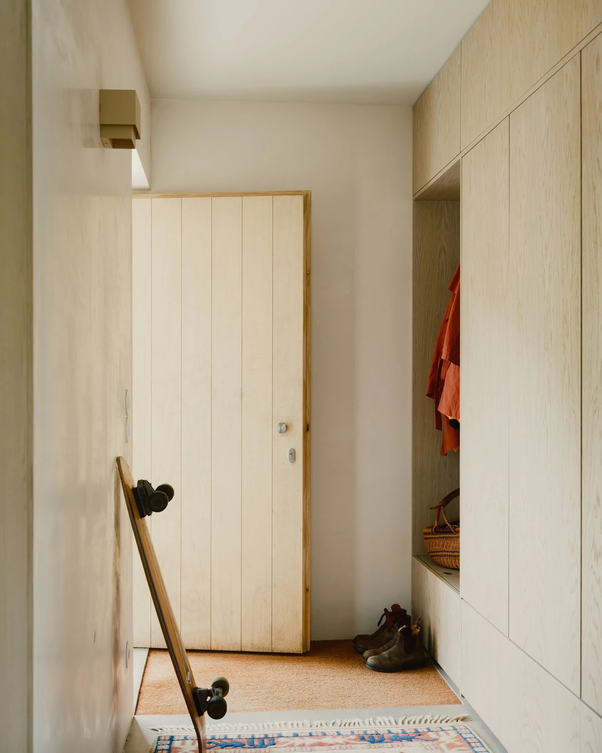Casa de Souvinha by Parto Atelier, photographed by Francisco Ascensão, Parto Atelier — The entry lines a wall of blond plywood joinery and a plank door against an orange terrazzo floor, a skateboard and worn boots left by the threshold.