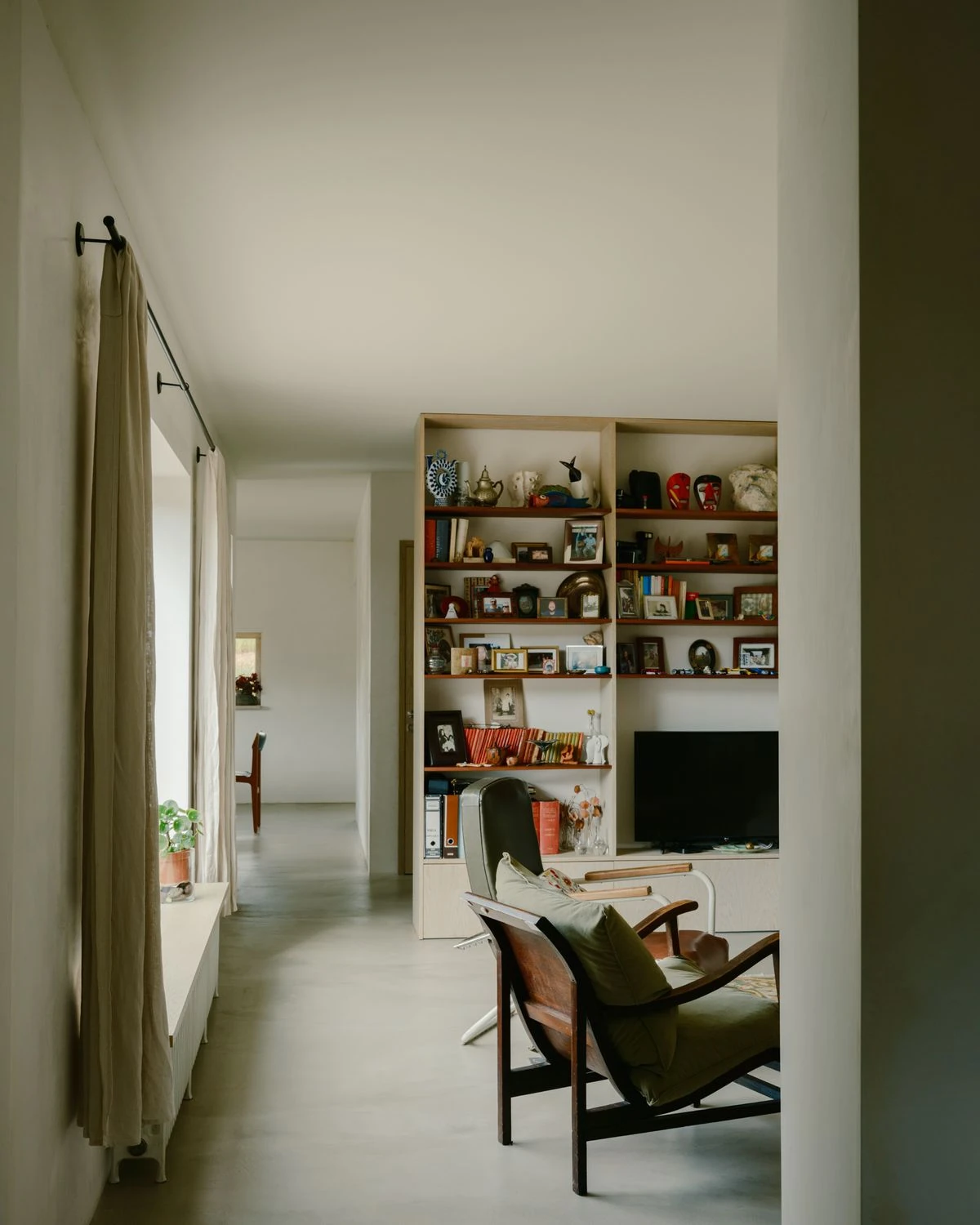 Casa de Souvinha by Parto Atelier, photographed by Francisco Ascensão, Parto Atelier — A hallway runs past built-in bookshelves crowded with objects and a mid-century leather chair, linen curtains softening the light over the microcement floor.