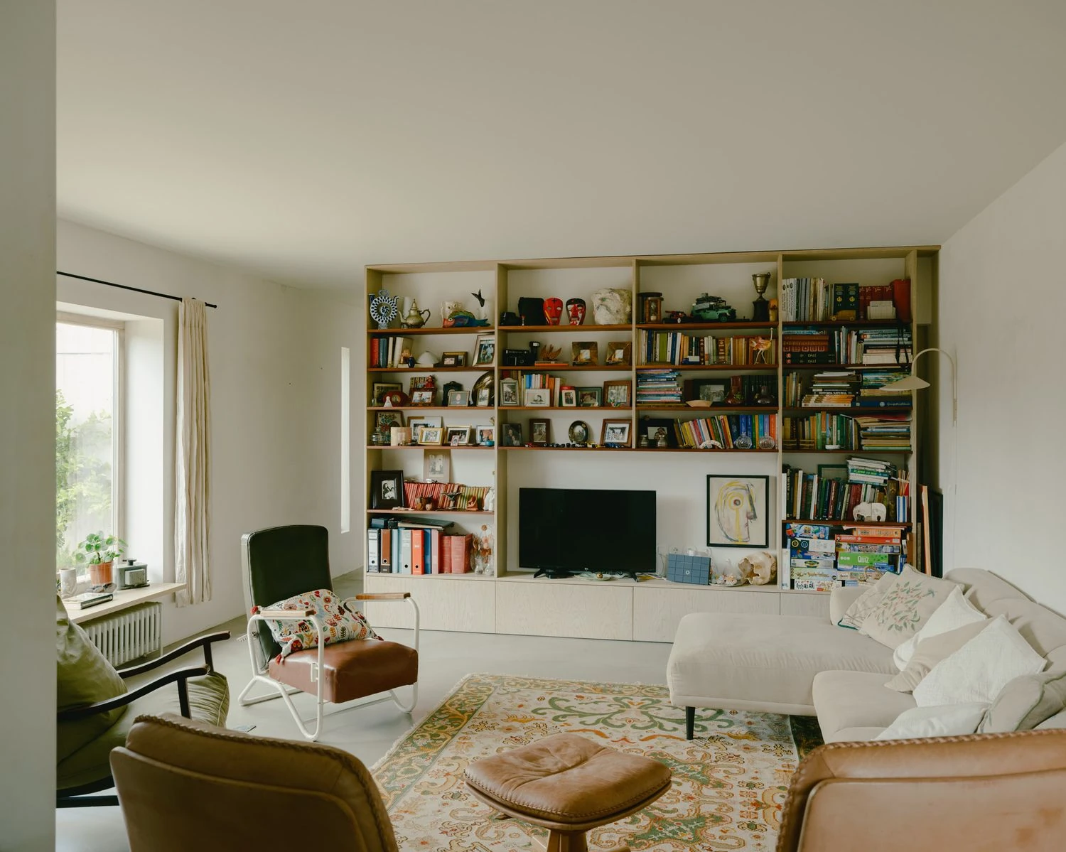 Casa de Souvinha by Parto Atelier, photographed by Francisco Ascensão, Parto Atelier — The living room fills a wall with built-in shelving of books and objects above a low cabinet, leather chairs and a patterned rug gathered on the pale floor.