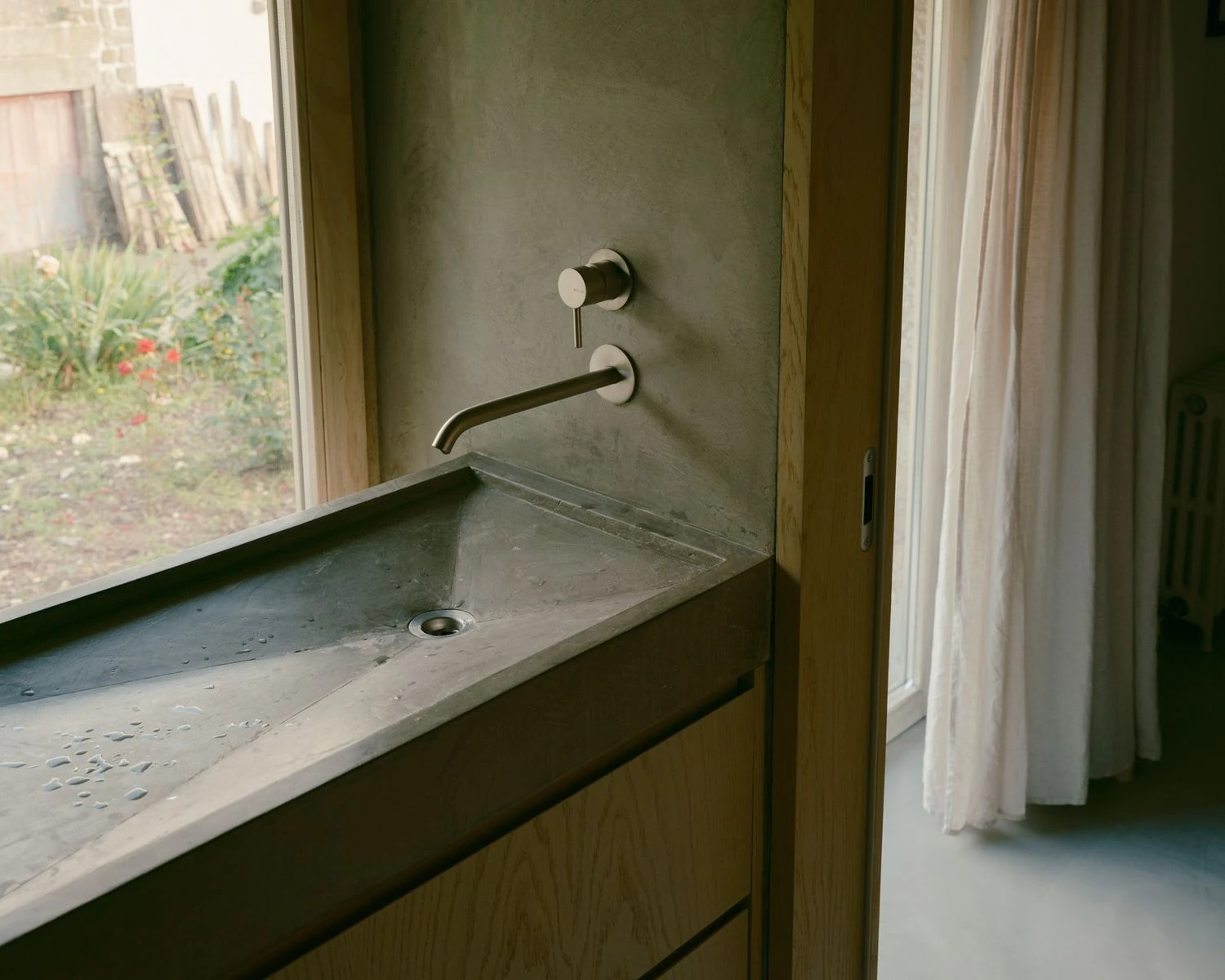 Casa de Souvinha by Parto Atelier, photographed by Francisco Ascensão, Parto Atelier — A cast concrete trough sink runs beneath a bedroom window with a brushed steel wall tap, the timber cabinet below opening to a view of the old stone yard.