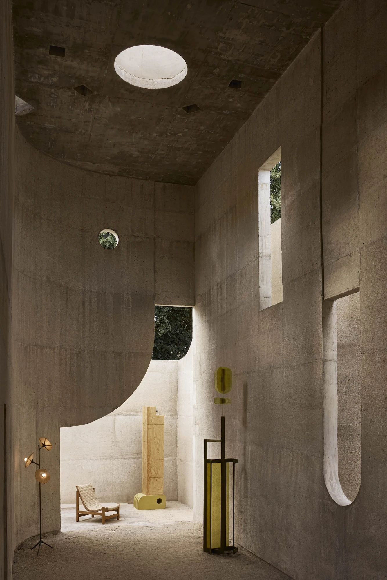 Casa Ínsula by TÁNAT, photographed by Alejandro Ramírez — A circular skylight punched through the roof slab drops daylight into a tall concrete room, where vertical slots and a round porthole open onto trees behind a plywood tower and brass floor lamp.