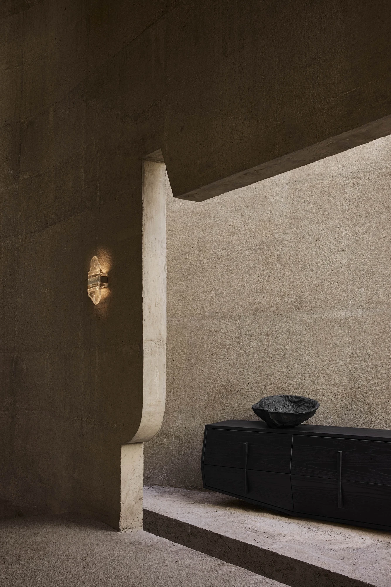 Casa Ínsula by TÁNAT, photographed by Alejandro Ramírez — A rock-crystal sconce glows against dark board-formed concrete above a black-oak sideboard, a rough black bowl catching the last light near a curved cut in the wall.
