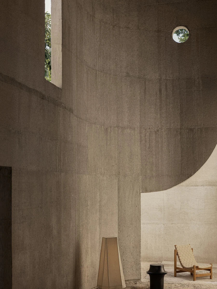 Casa Ínsula by TÁNAT, photographed by Alejandro Ramírez — A tall concrete room lit through a narrow vertical slot and a small round porthole cut straight through the wall, with a woven leather chair and a fabric lamp set on the sand floor.