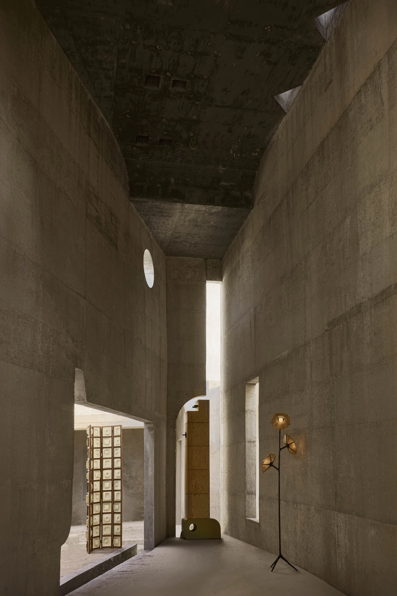 Casa Ínsula by TÁNAT, photographed by Alejandro Ramírez — A high concrete corridor lit from an oval oculus and a thin light slot, a folding tile screen and a plywood tower standing against the sandy walls.