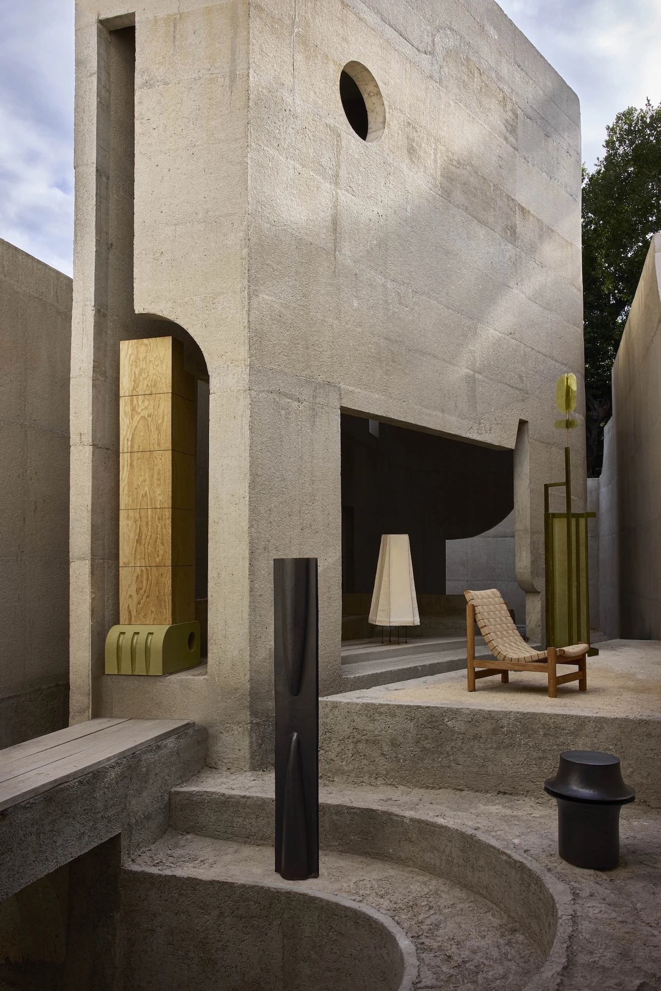 Casa Ínsula by TÁNAT, photographed by Alejandro Ramírez — An exterior corner of raw concrete pierced by a round oculus, with a plywood block set in an arched niche and dark objects arranged across broad concrete steps.