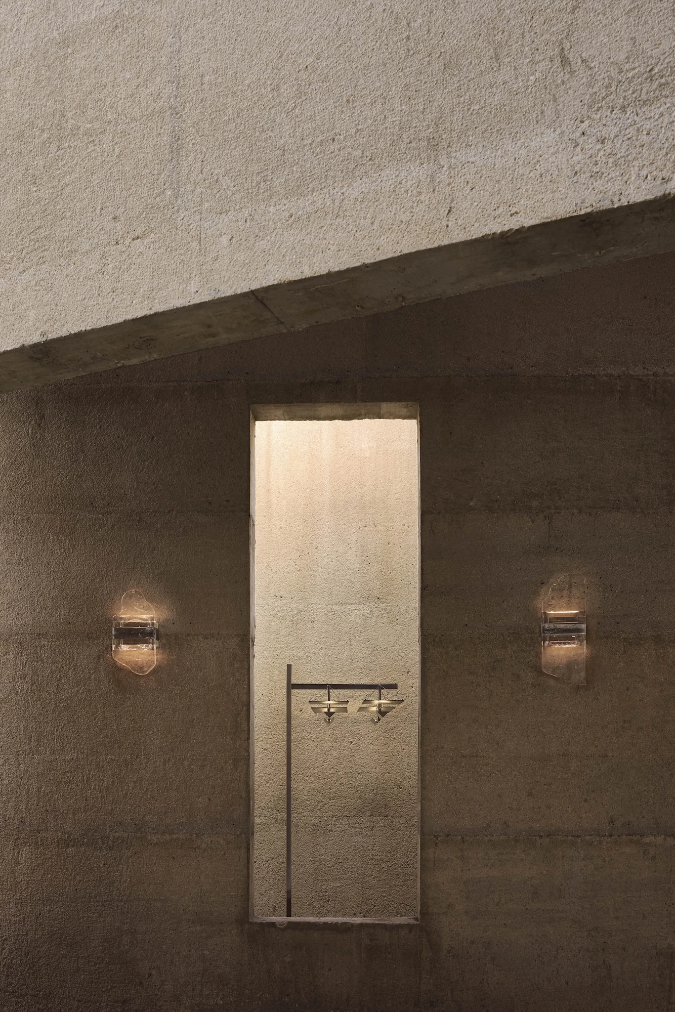 Casa Ínsula by TÁNAT, photographed by Alejandro Ramírez — Two rock-crystal sconces flank a deep vertical niche lit from within, brass fittings mounted on the board-formed concrete face.