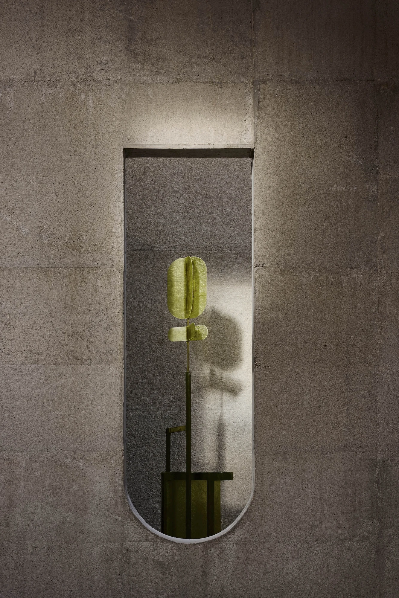 Casa Ínsula by TÁNAT, photographed by Alejandro Ramírez — An olive-green sculptural lamp stands lit inside a tall arched slot cut through thick concrete, its shadow thrown across the sandy wall.