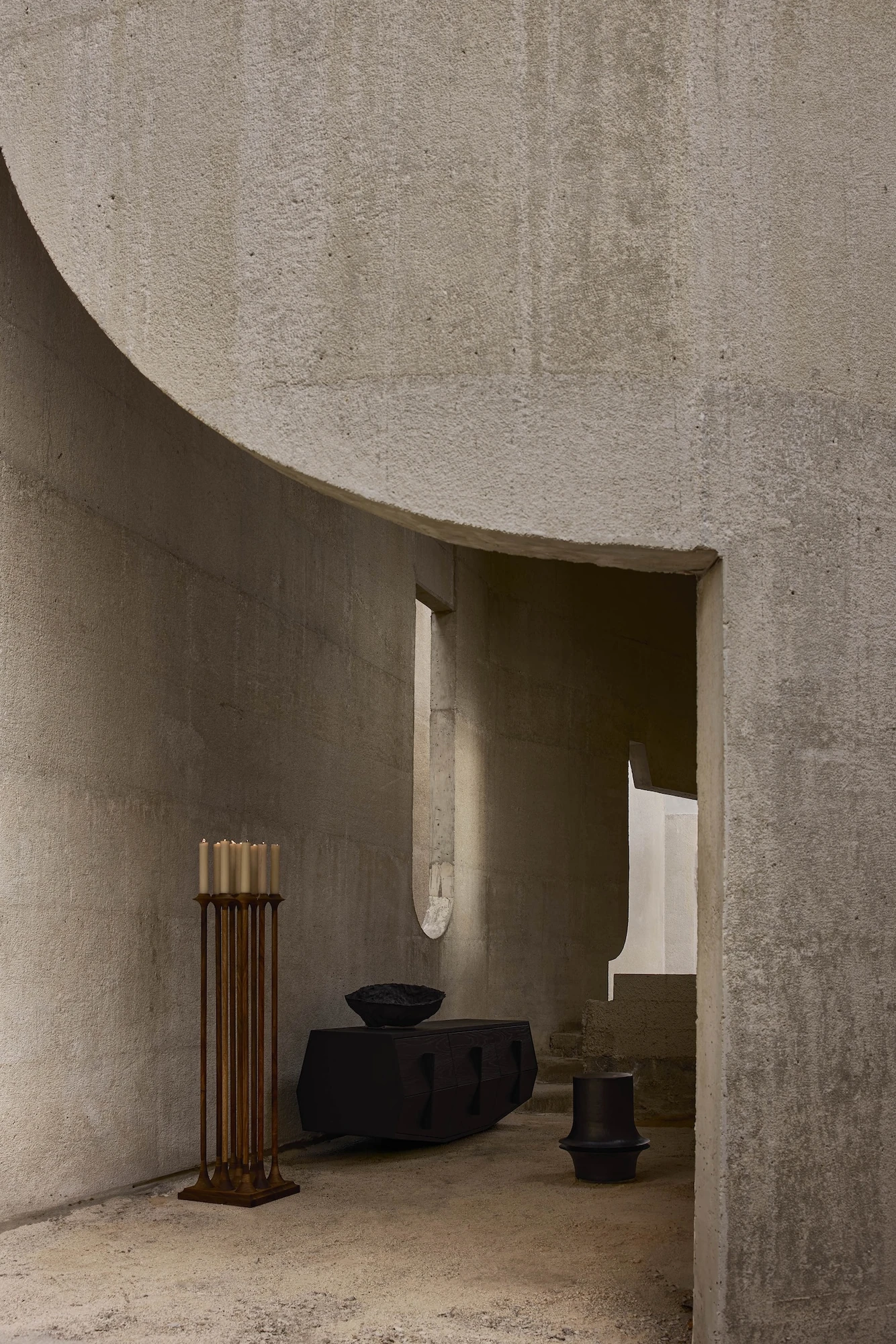 Casa Ínsula by TÁNAT, photographed by Alejandro Ramírez — A curved concrete soffit sweeps overhead above a tall candle stand, a black chest and dark vessels near a doorway that steps down toward daylight.