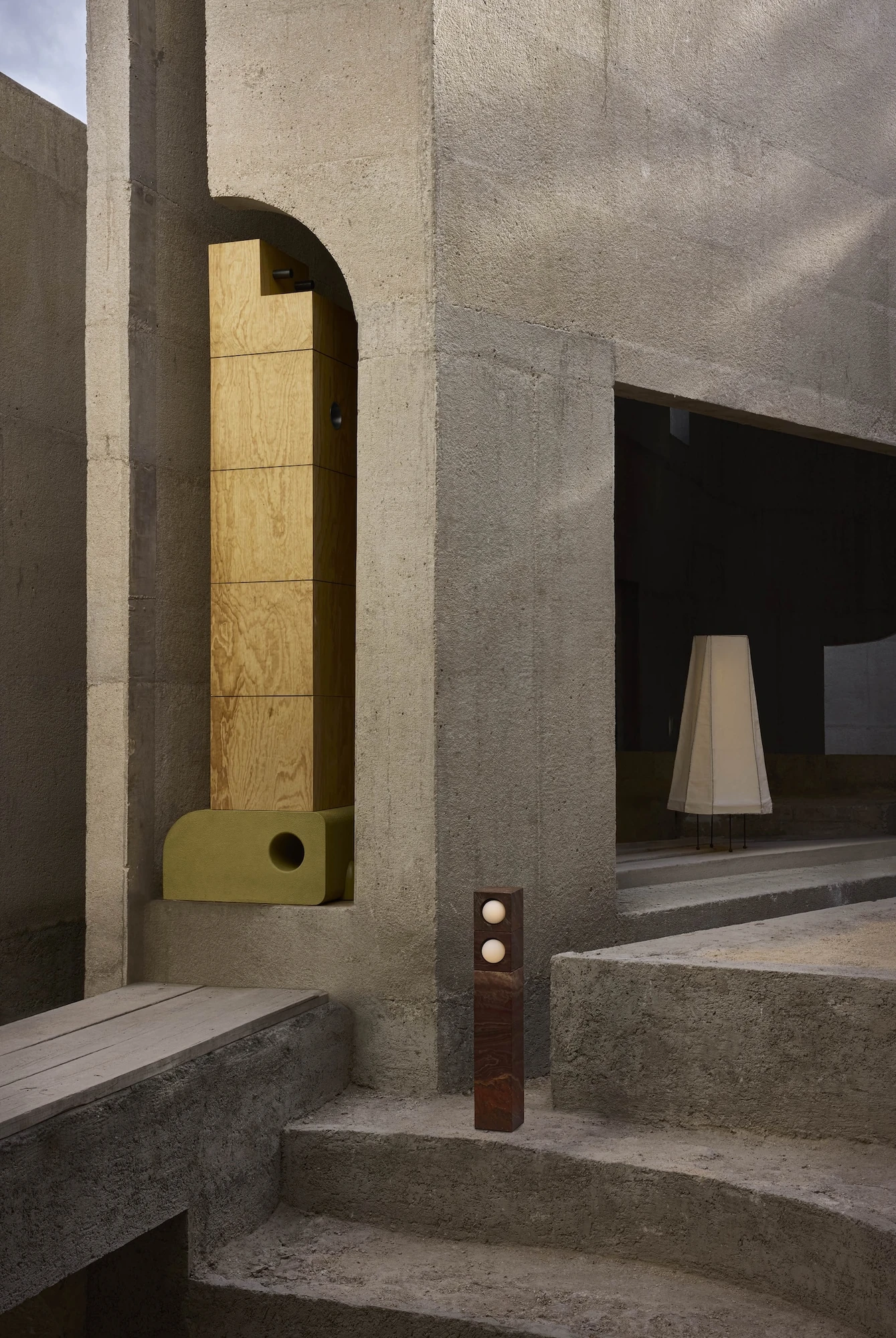 Casa Ínsula by TÁNAT, photographed by Alejandro Ramírez — An exterior arched niche holds a plywood tower on an olive base, a stacked-sphere lamp and a fabric lamp glowing in the dark opening beyond the concrete steps.