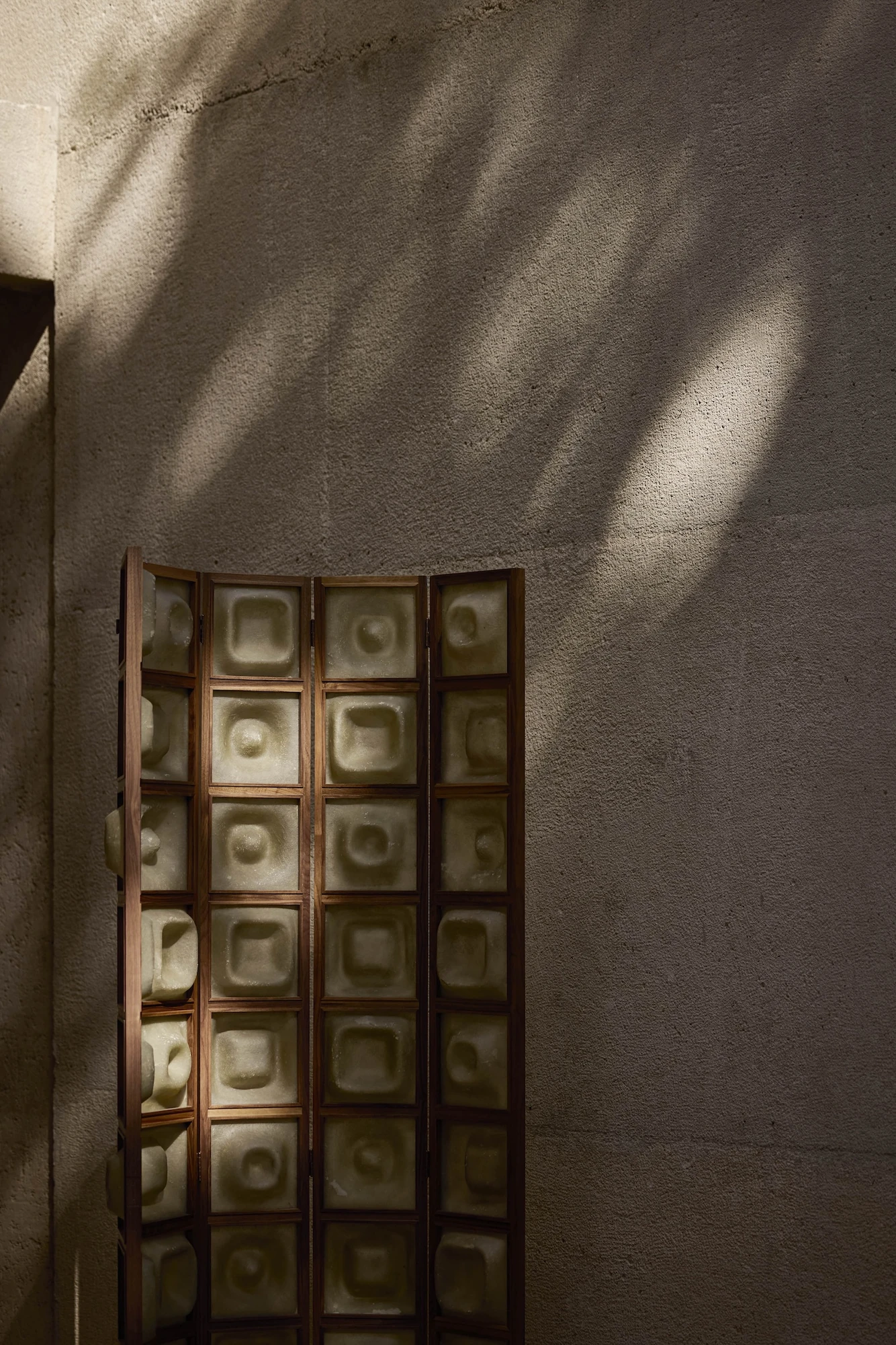 Casa Ínsula by TÁNAT, photographed by Alejandro Ramírez — A folding screen of pale green cast tiles in a wooden frame stands against board-formed concrete raked by dappled daylight.