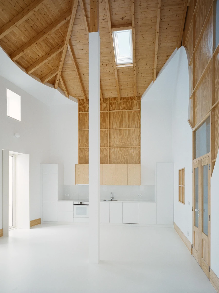 Cutouts by Corpo Atelier, photographed by Francisco Ascensão — A double-height white room where a slender white pillar rises to a hipped timber roof, a skylight set into its pine slope above a flush white kitchen.