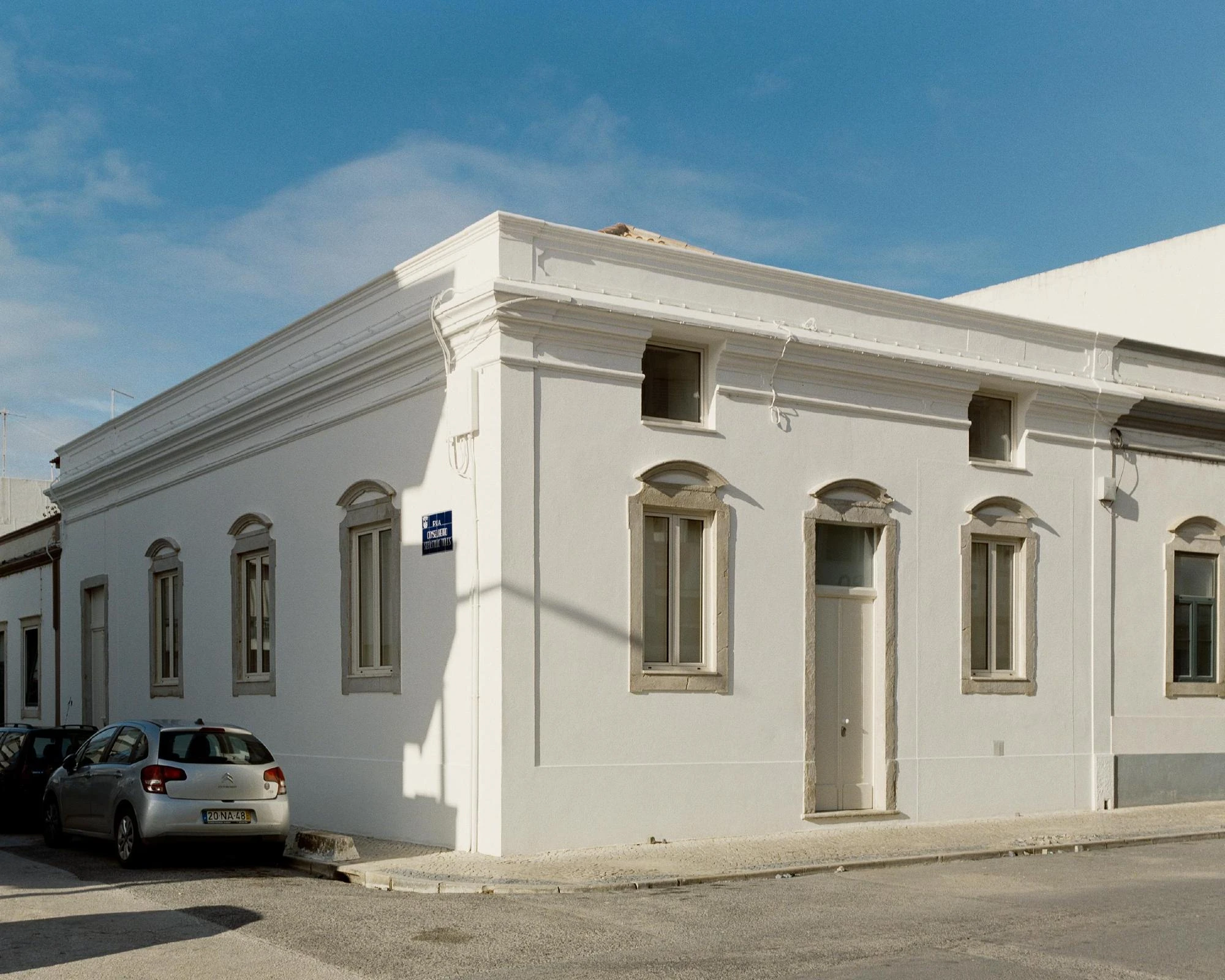 Cutouts by Corpo Atelier, photographed by Francisco Ascensão — The white plastered corner façade of the existing house, arched stone window surrounds and a moulded cornice under a clear blue sky, a silver car parked at the kerb.