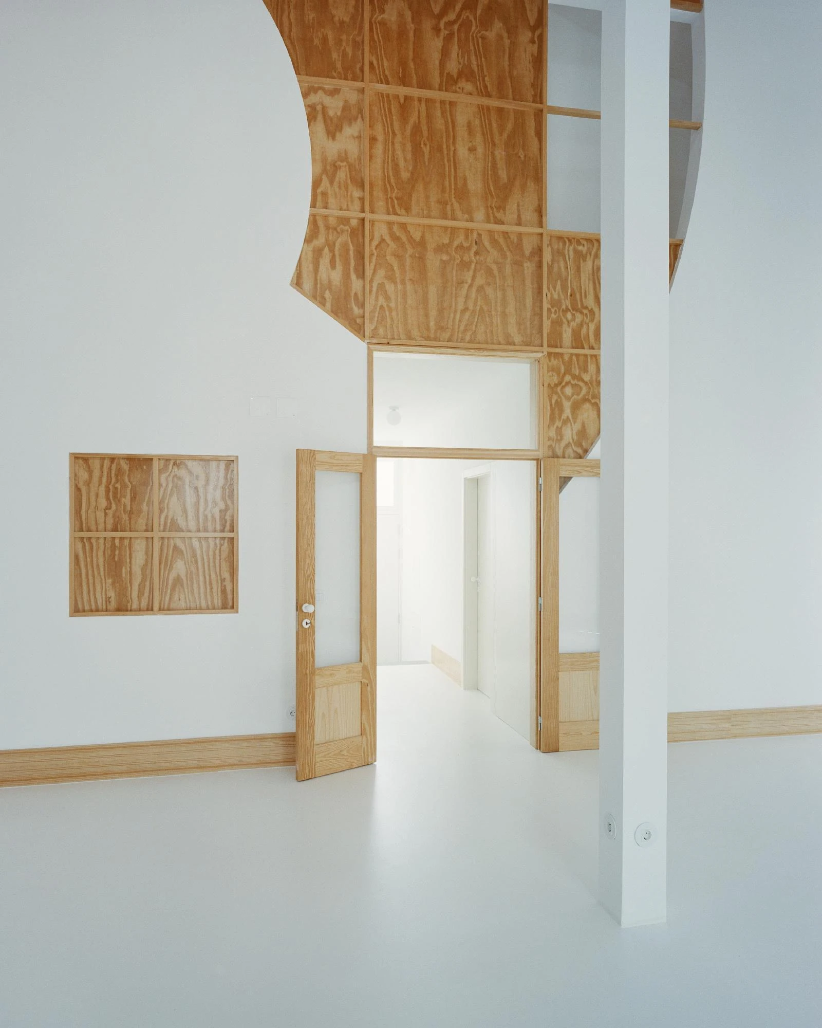 Cutouts by Corpo Atelier, photographed by Francisco Ascensão — A large organically curved cut opens a white wall to reveal maritime pine plywood behind it, a glazed pine door swung open onto a bright corridor.