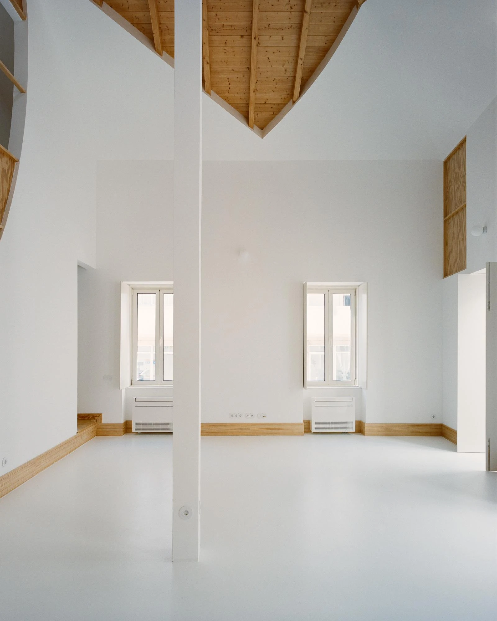 Cutouts by Corpo Atelier, photographed by Francisco Ascensão — A white pillar splits the view of two tall windows in a double-height room, pine skirting running low along the wall as the timber roof folds down at the top.