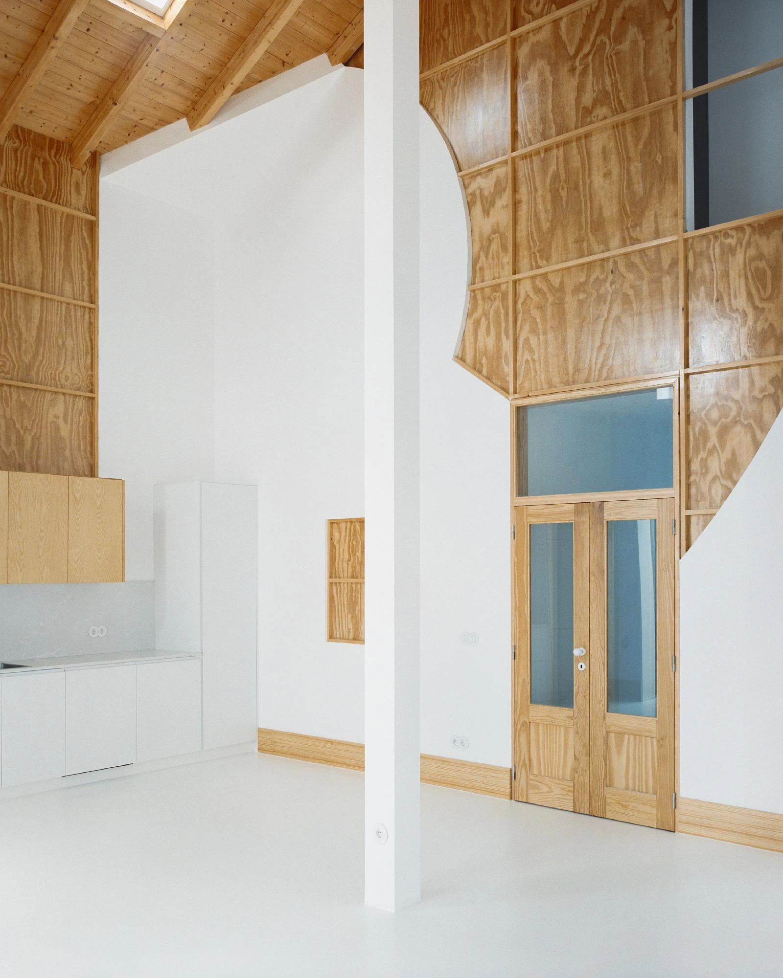 Cutouts by Corpo Atelier, photographed by Francisco Ascensão — A curved cut through plywood cladding frames a glazed pine double door with pale blue glass, a white pillar dividing the room from the kitchen beyond.