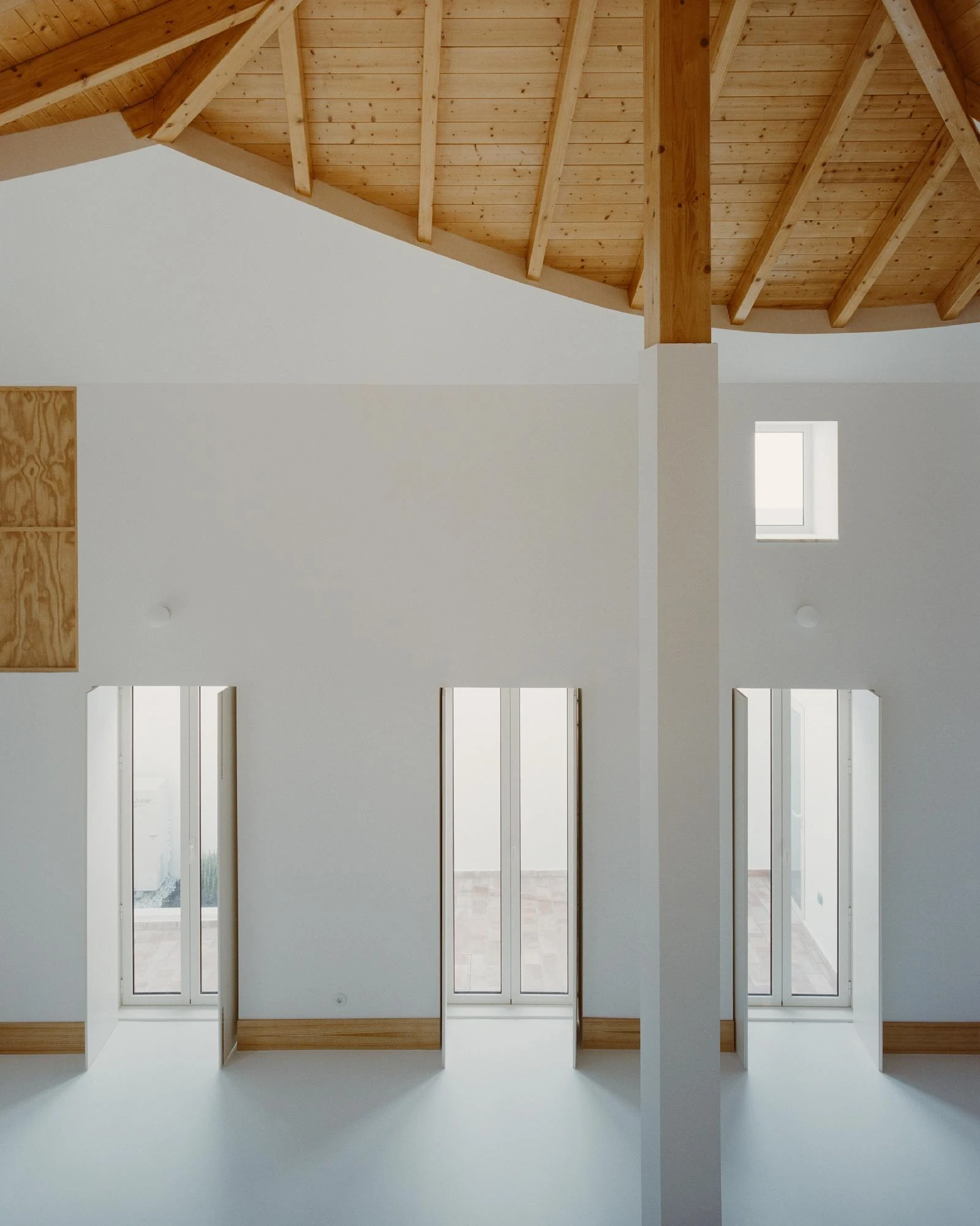 Cutouts by Corpo Atelier, photographed by Francisco Ascensão — Three tall narrow window reveals cut down to a terracotta-paved terrace, a white pillar meeting the pine roof, a plywood panel set high on the white wall.