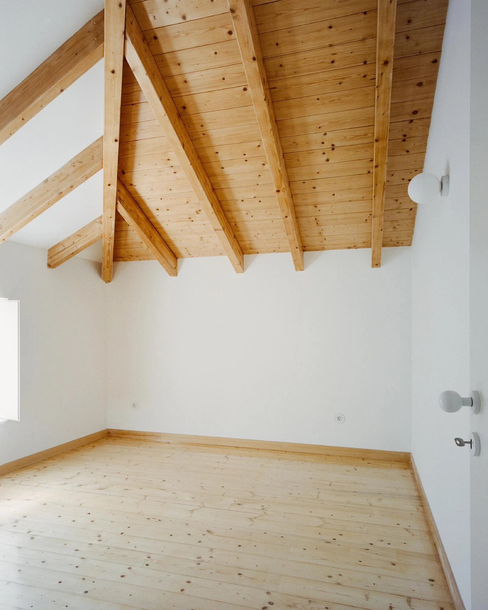 Cutouts by Corpo Atelier, photographed by Francisco Ascensão — An attic room lined in pale pine, exposed rafters overhead and plank flooring below, white walls and small round white door hardware.