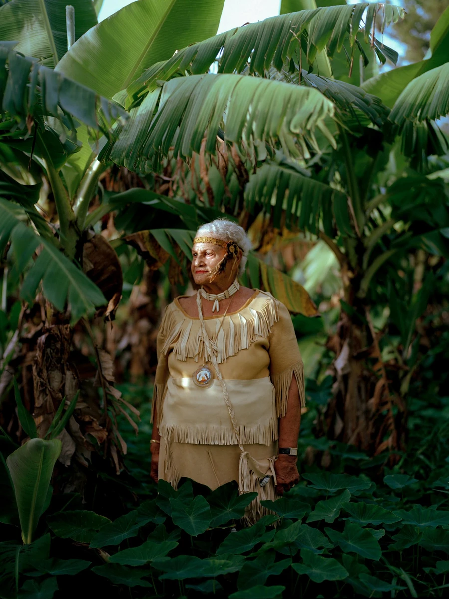 Descendants of Summer by Nicola Muirhead, photographed by Nicola Muirhead — An older woman in a fringed tan buckskin dress and beaded medallion stands among broad banana leaves, her white hair catching the afternoon light.