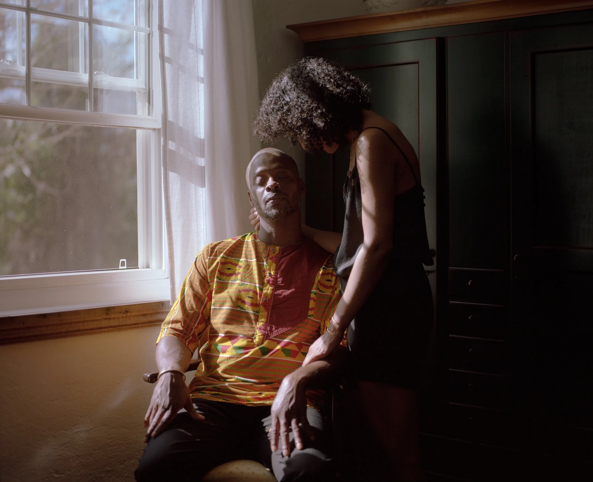 Descendants of Summer by Nicola Muirhead, photographed by Nicola Muirhead — In a domestic interior, a seated man in a kente-cloth shirt closes his eyes as a woman in a black slip stands behind him, her hand at his neck, window light falling across them…