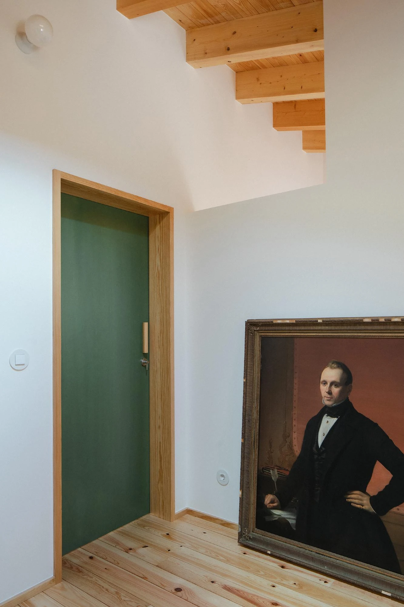 House in Cerrado by Atelier Espaço P2, photographed by José Campos — A forest-green door in a pale pine frame stands beside an antique oil portrait leaning against a white wall under exposed beams.