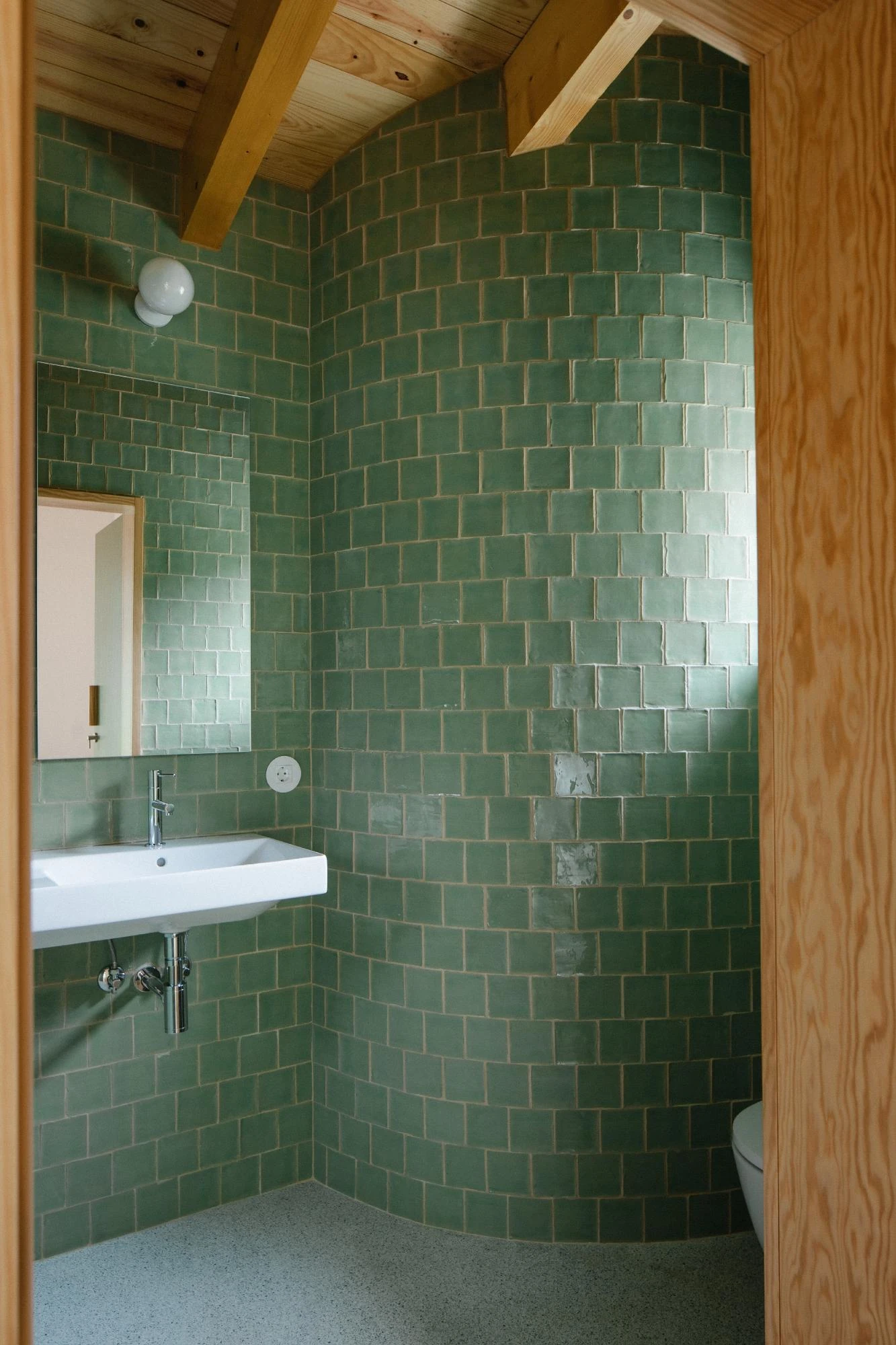 House in Cerrado by Atelier Espaço P2, photographed by José Campos — A washroom is lined floor to ceiling in hand-glazed square green tile, curving around a corner past a white basin and mirror beneath a pine ceiling.