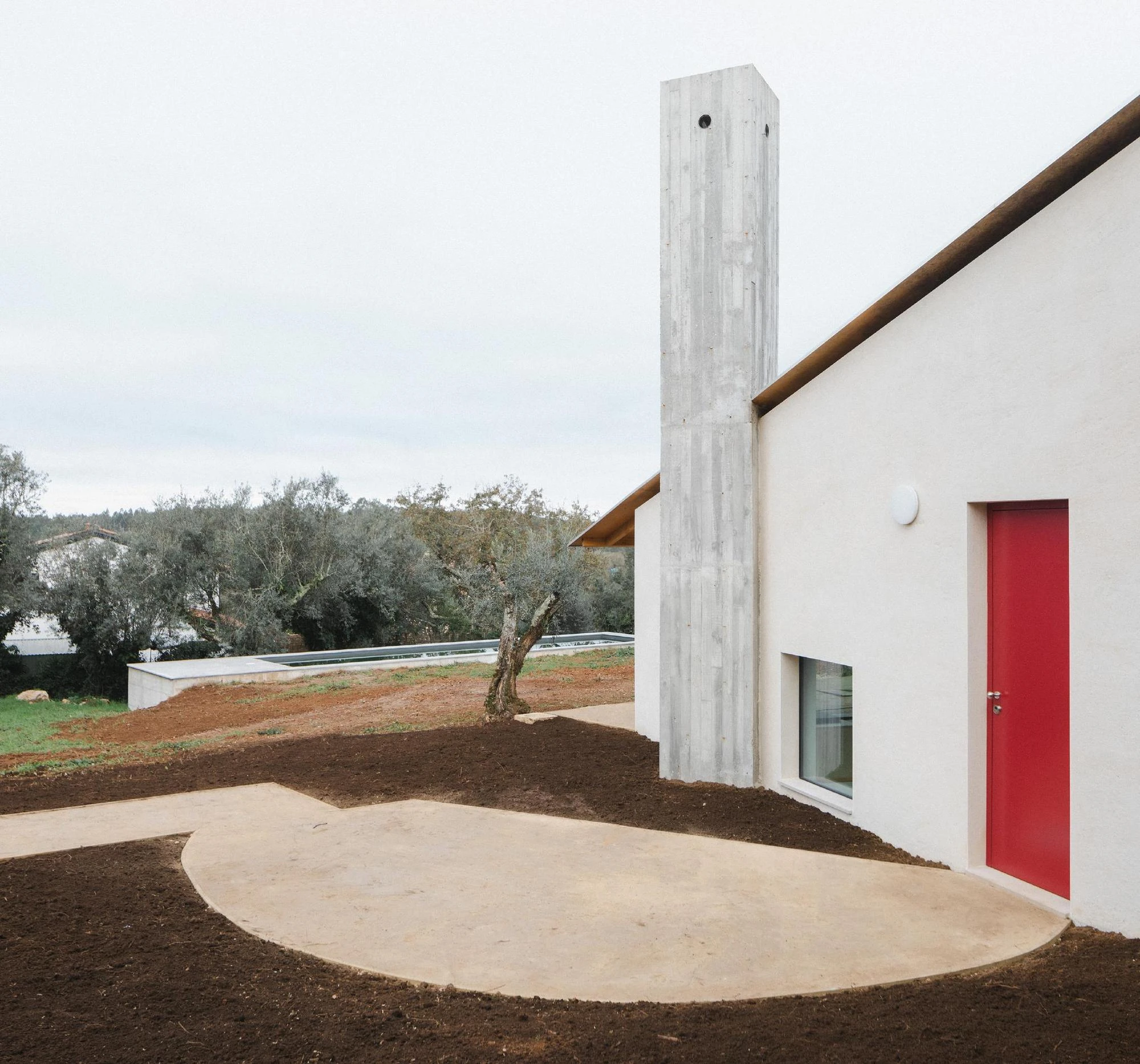 House in Cerrado by Atelier Espaço P2, photographed by José Campos — The full-height board-formed concrete chimney rises beside a white wall and red door, a curved concrete path sweeping past toward a lap pool in the distance.