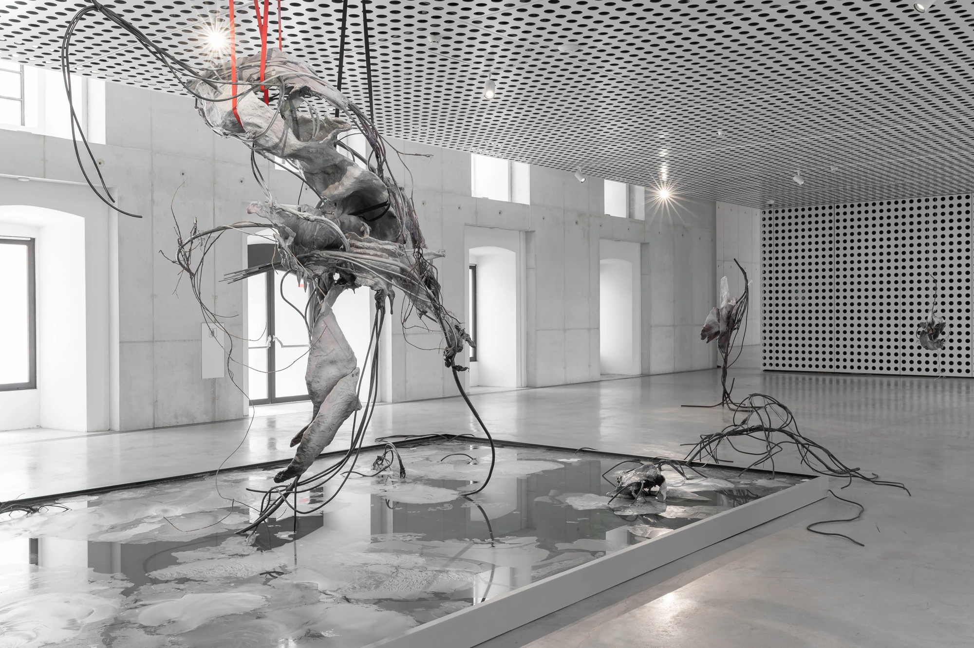 Accomplice by Yein Lee, photographed by Blaž Gutman / MGML — One of Lee's suspended sculptures tips out over the ground-based pool, pale cast masses tangled with dark cabling and red straps.