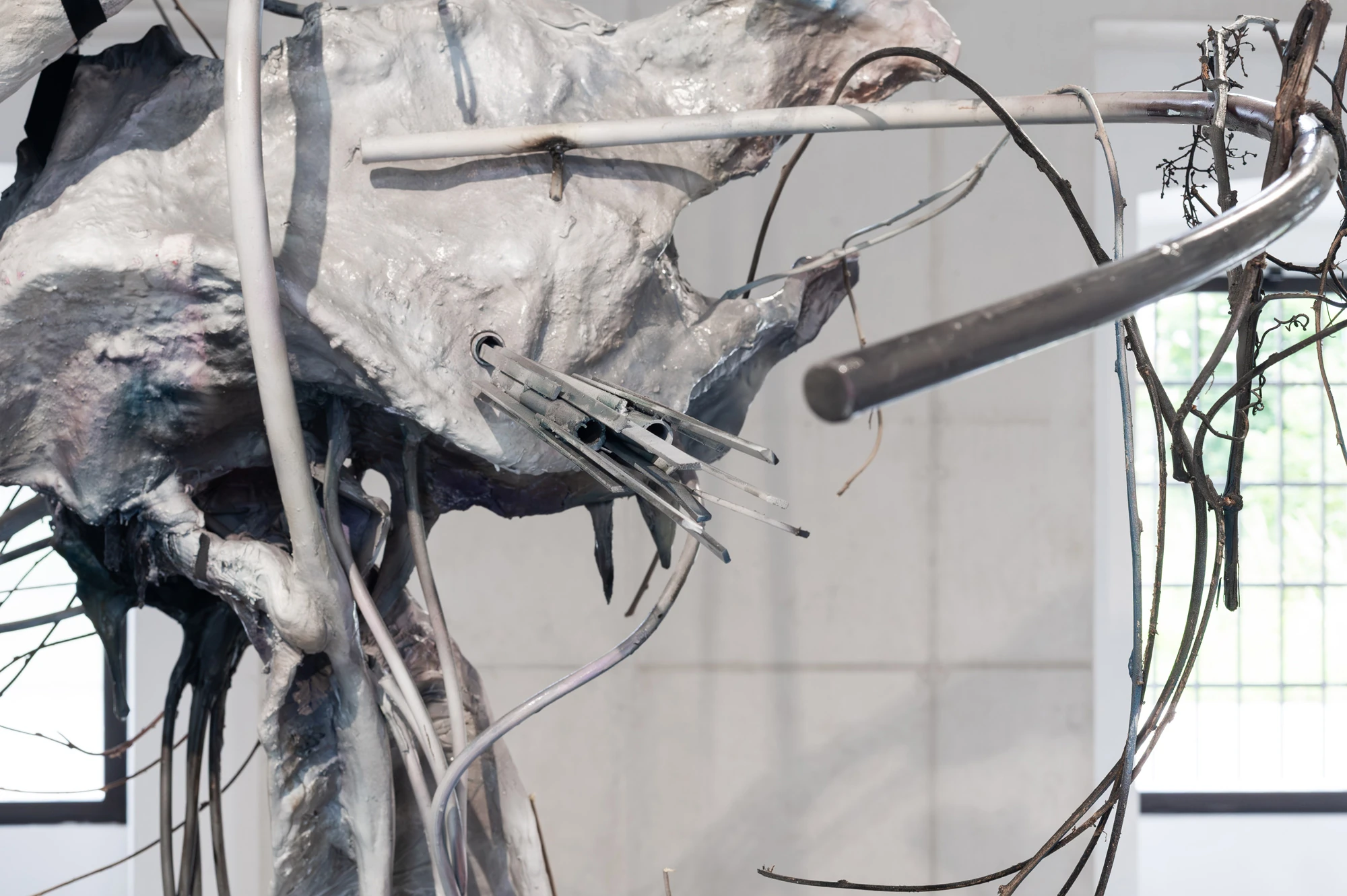 Accomplice by Yein Lee, photographed by Blaž Gutman / MGML — Close view of a sculpture in Accomplice, a pale cast mass pierced by a white curved tube, a cut grey rod and dry branches.