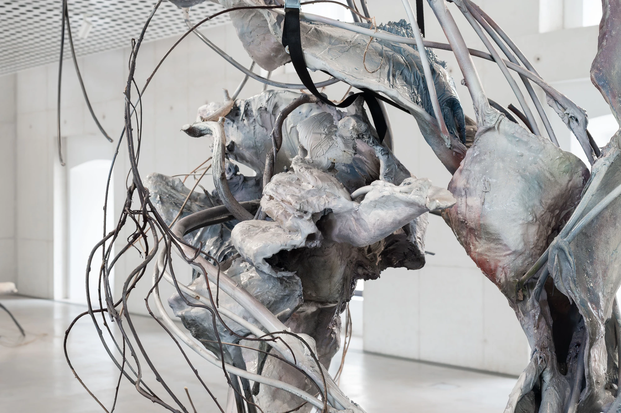 Accomplice by Yein Lee, photographed by Blaž Gutman / MGML — Detail of a sculpture, blue-grey and white cast folds threaded with pale tubing and thin branches inside the concrete gallery.