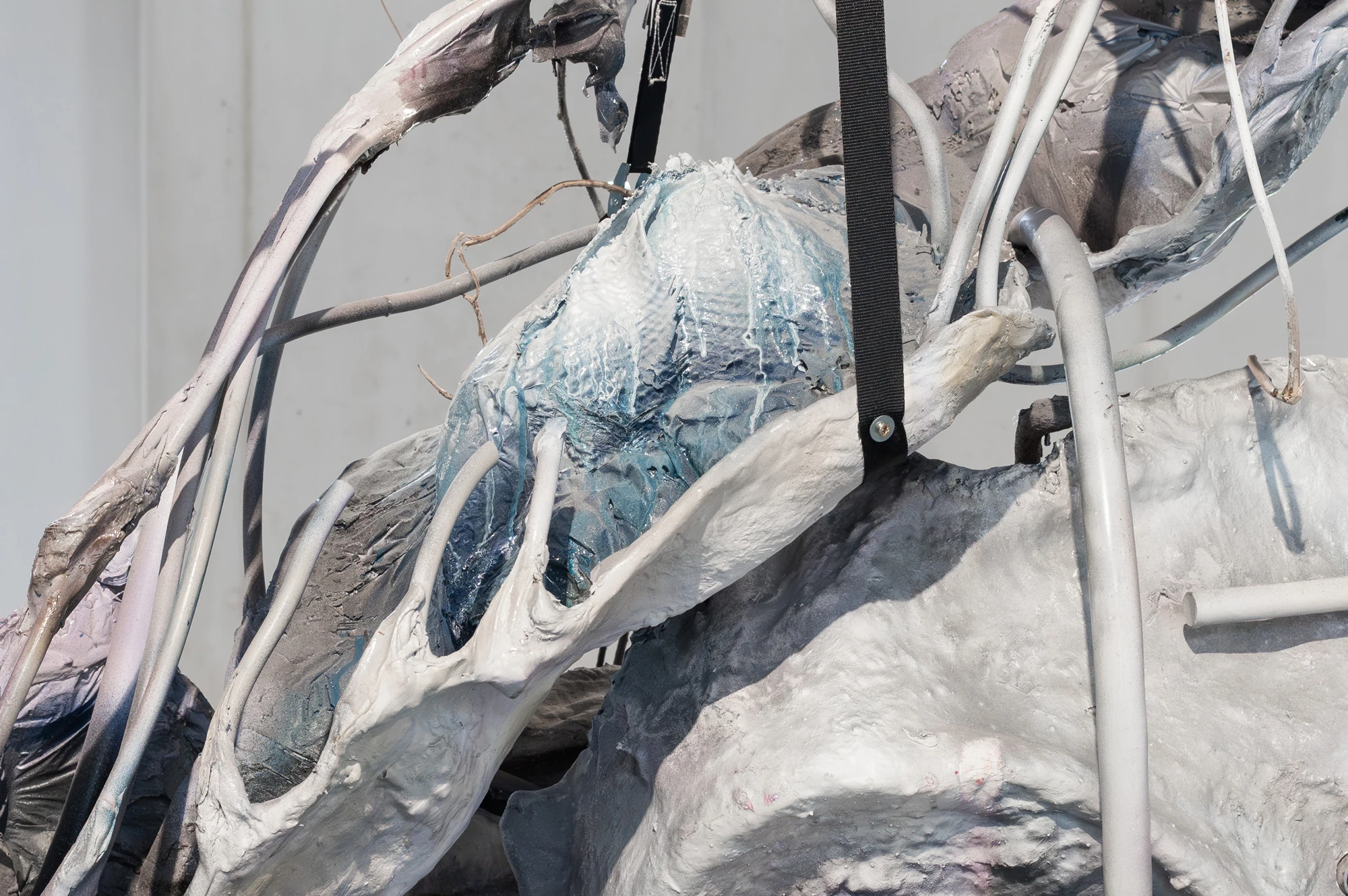 Accomplice by Yein Lee, photographed by Blaž Gutman / MGML — Close detail of a sculpture, blue and white cast surfaces held together with a black strap and grey tubing.