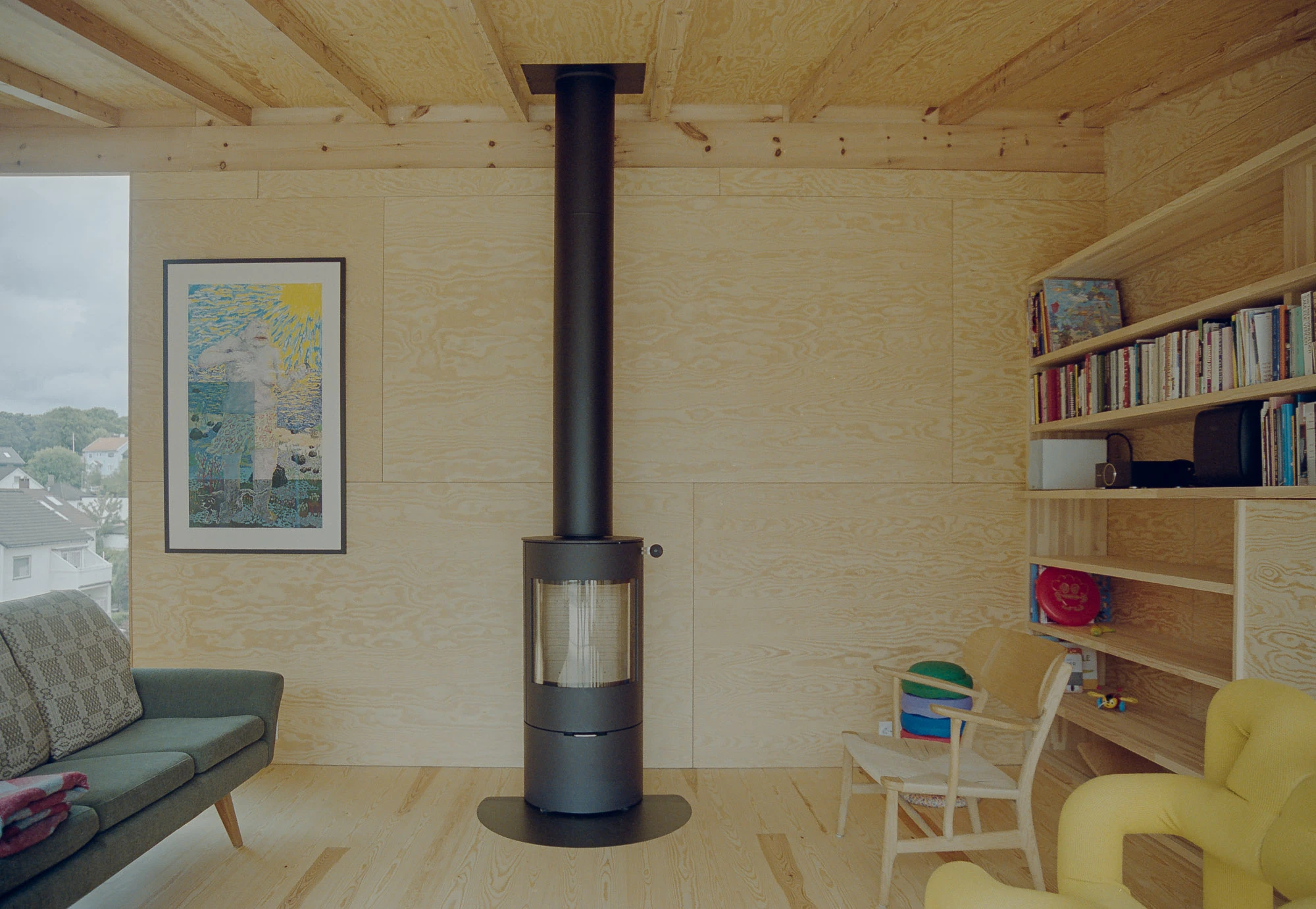 Ersland & Skeie House by Knut Hjeltnes Architects, photographed by Hugo Lütcherath — Peeled pine plywood walls carry built-in bookshelves beside a black wood-burning stove and a low sofa.